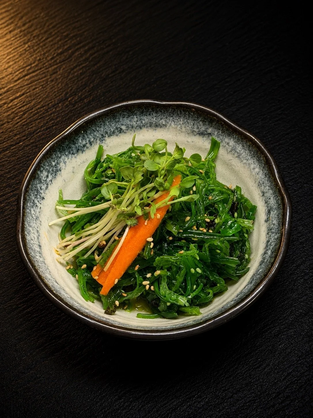 A bowl of seaweed salad topped with microgreens, shredded carrots, and sesame seeds on a black surface.