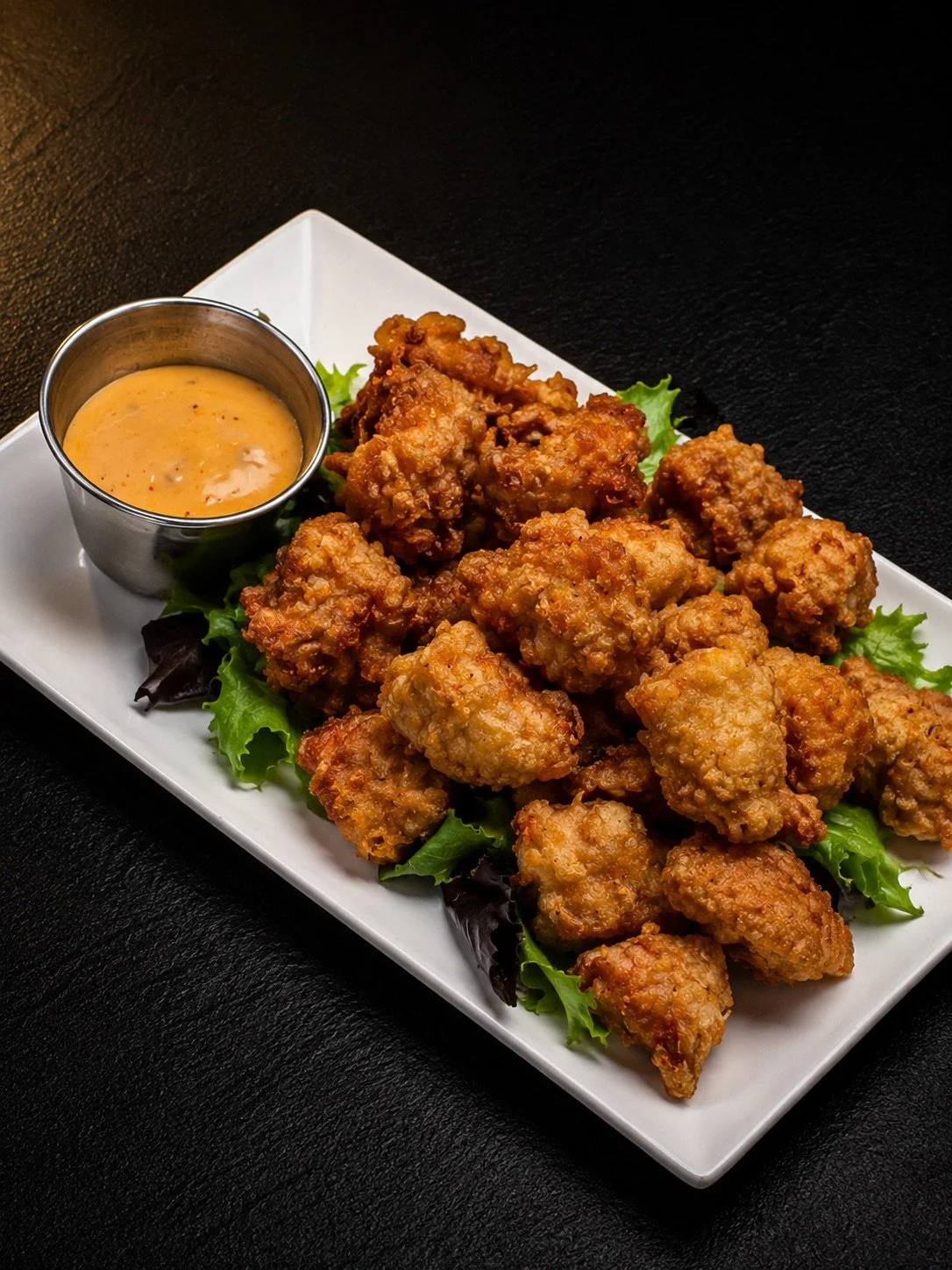 Plate of fried chicken pieces with a side of dipping sauce on a rectangular white dish, garnished with leafy greens, on a black surface.