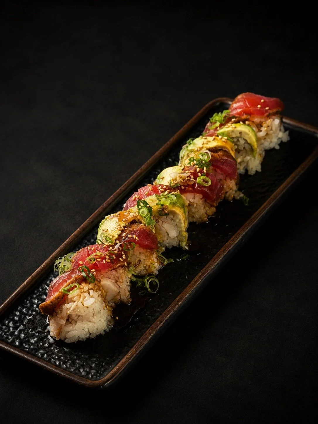 Sushi roll with rice, raw fish, avocado, and green onions, garnished with sesame seeds on a black rectangular plate.