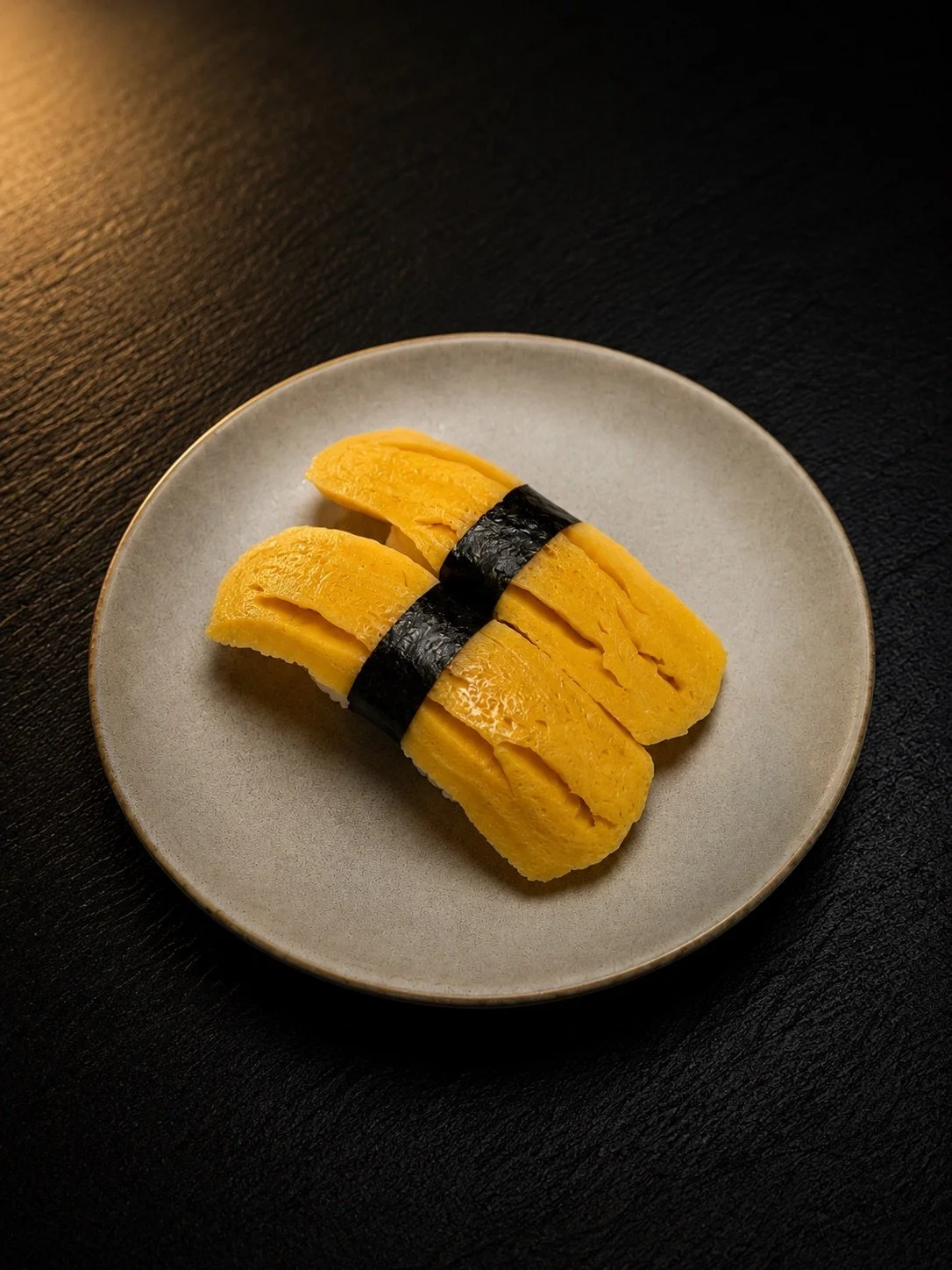 A plate with two pieces of tamago sushi with sliced yellow egg omelette on top, held together with a strip of black seaweed, on a black textured surface.