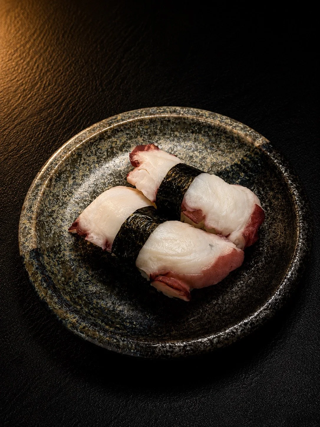 Two pieces of sushi with white fish and seaweed on a textured black plate.