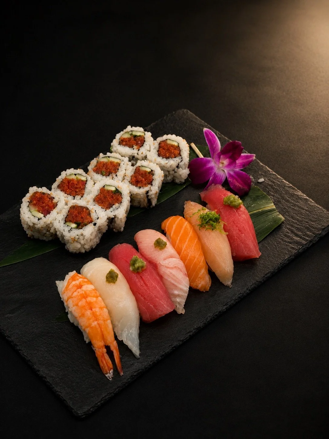 Assorted sushi platter with rolls, nigiri, a purple orchid, and green garnish on a black slate serving tray.