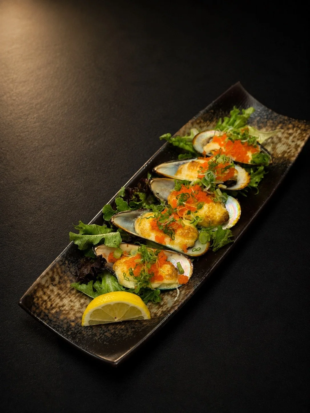 A rectangular black ceramic plate with five cooked oysters on a bed of greens, garnished with chopped green onions, orange fish roe, and a lemon wedge, set against a dark background.