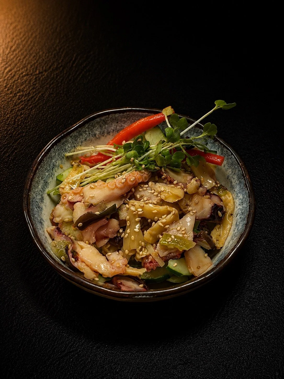 A bowl of mixed Asian-style vegetables topped with sesame seeds, garnished with microgreens and red peppers, set against a black background.