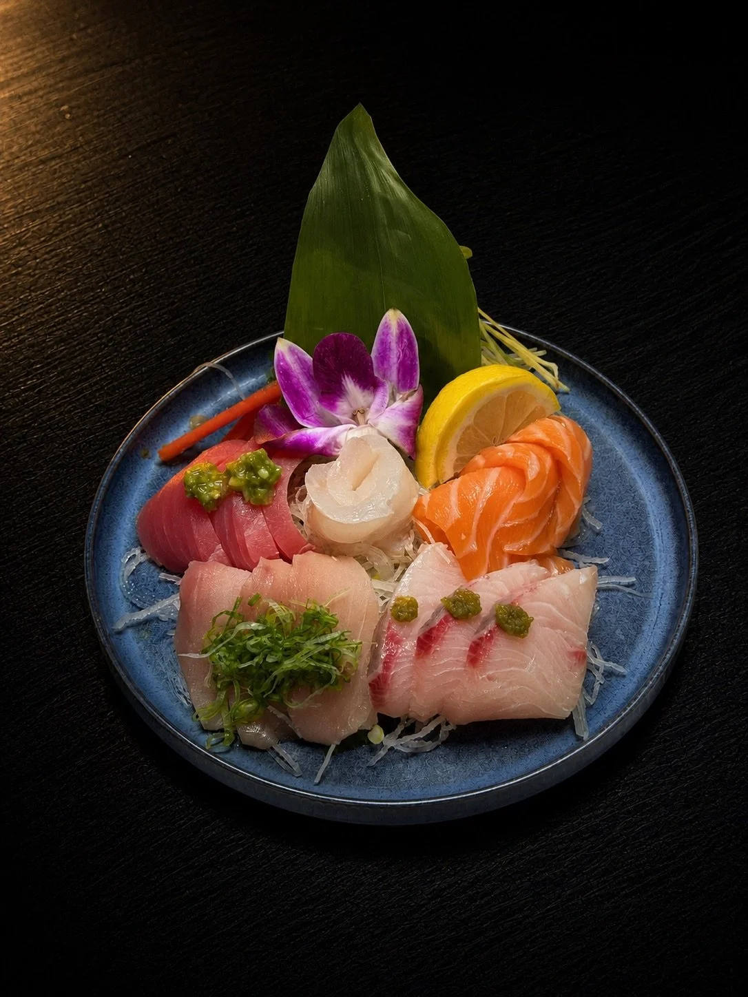 Assorted sashimi platter with slices of raw fish, garnished with wasabi, lemon, edible flowers, greens, and a leaf, served on a dark table