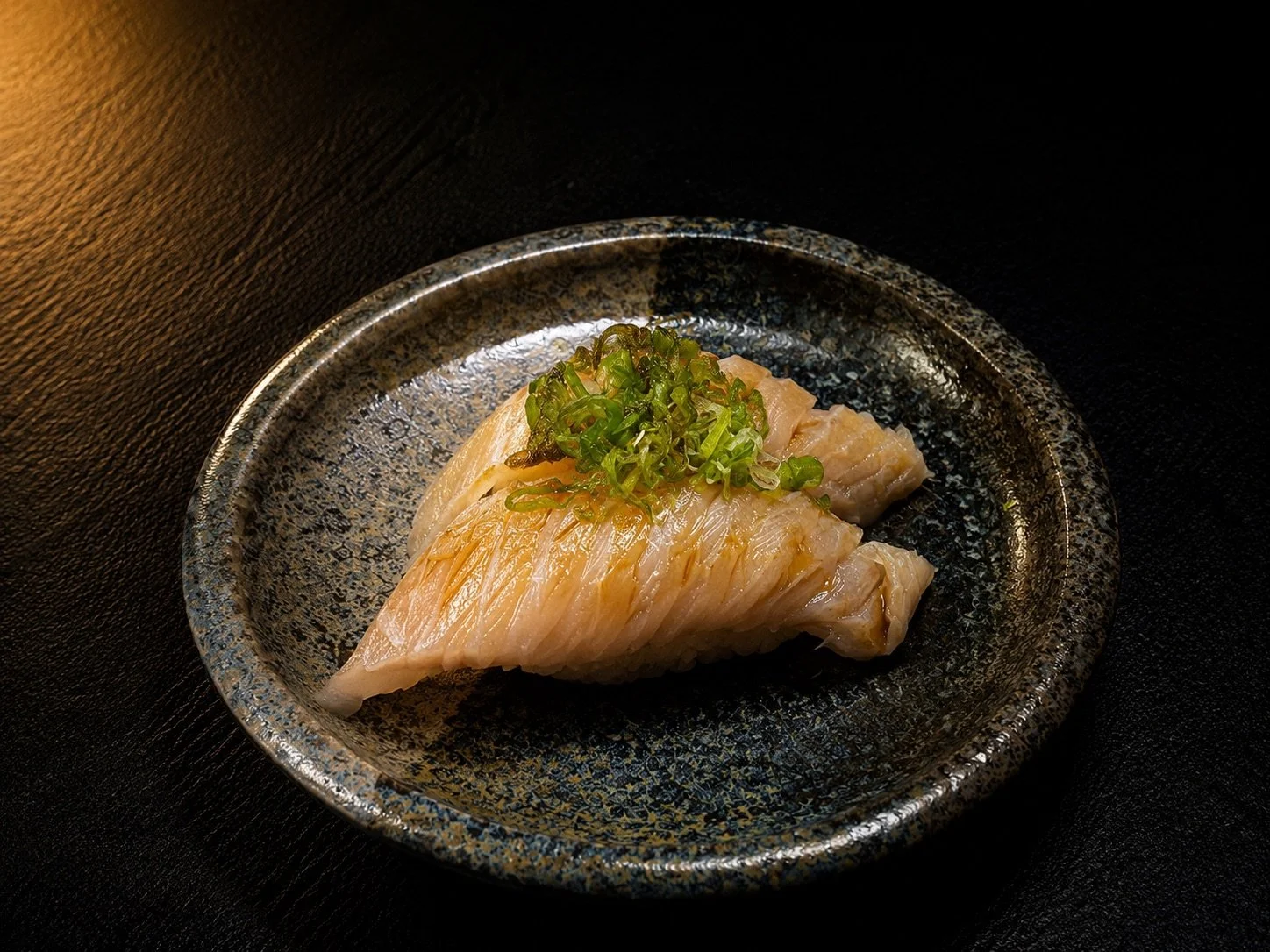 A piece of cooked fish topped with chopped green onions on a black ceramic plate.