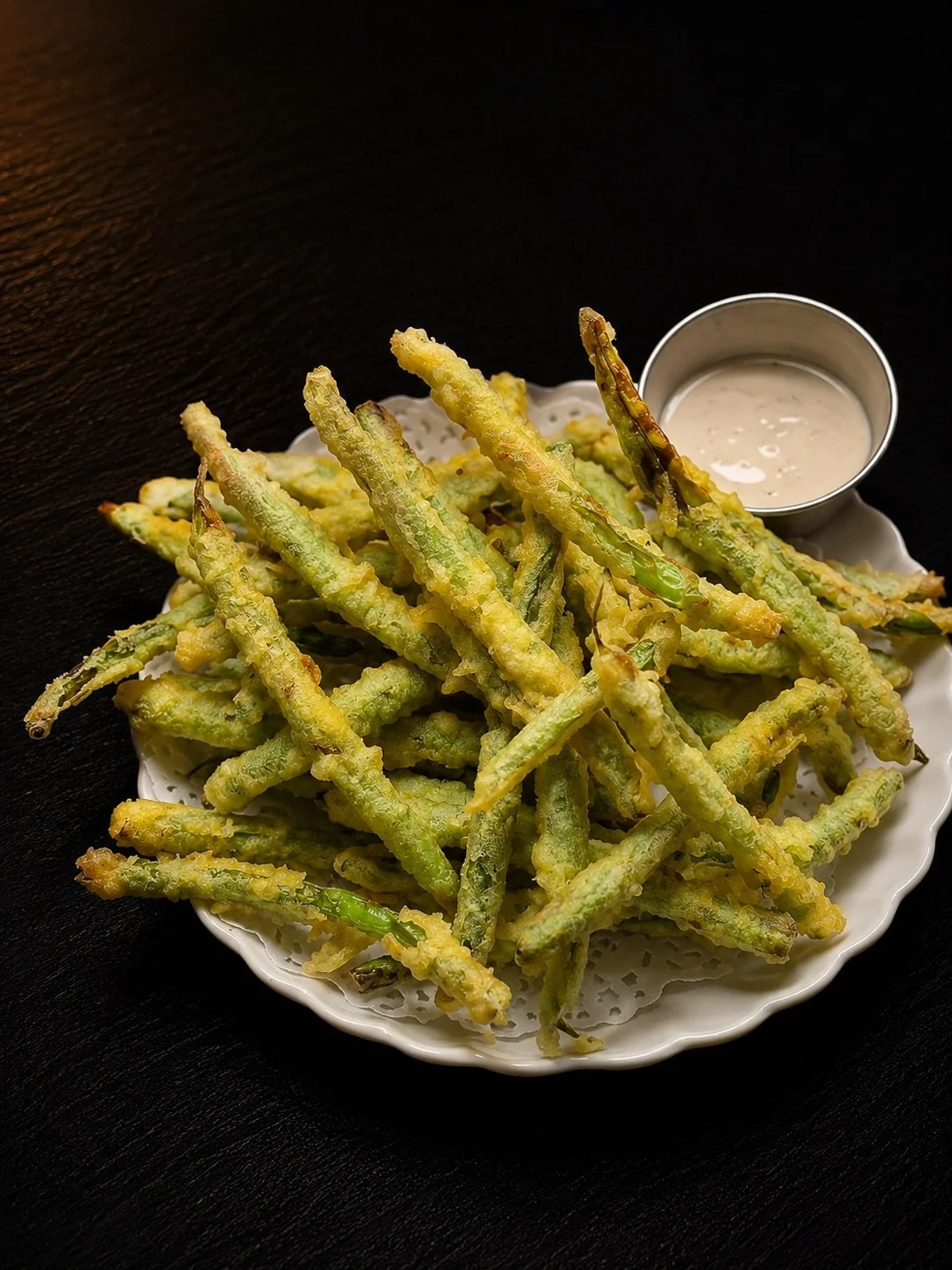 Bowl of tempura green beans with dipping sauce on the side, placed on a dark wooden surface.