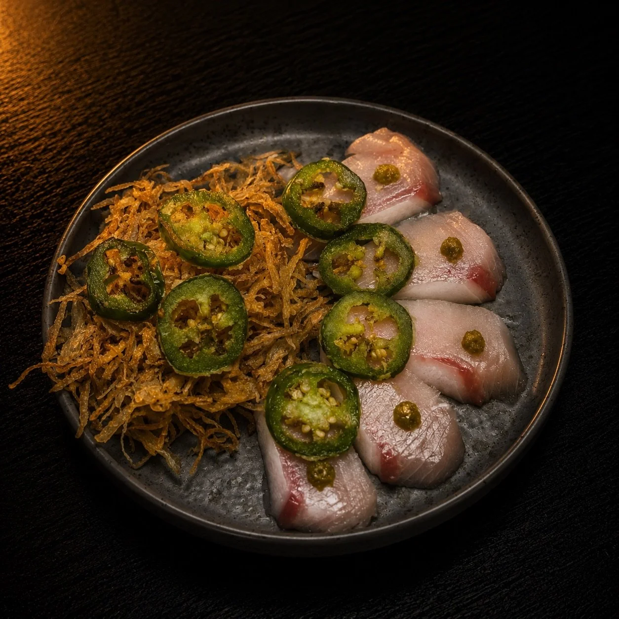Sliced fish on a dark plate garnished with sliced jalapeños and green sauce, served with shredded fried onions.