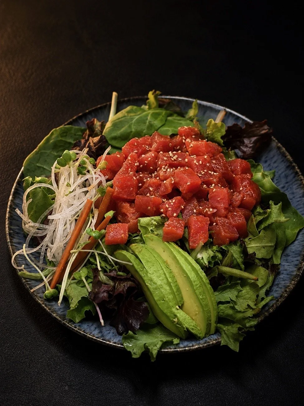 Salad with mixed greens, avocado slices, diced raw fish or tuna, shredded radish, and sesame seeds on a black background.