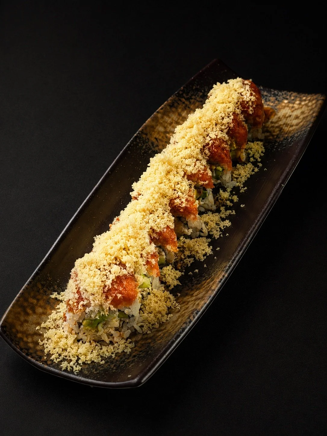 Sushi roll topped with shredded cheese and spicy sauce on a black rectangular plate.