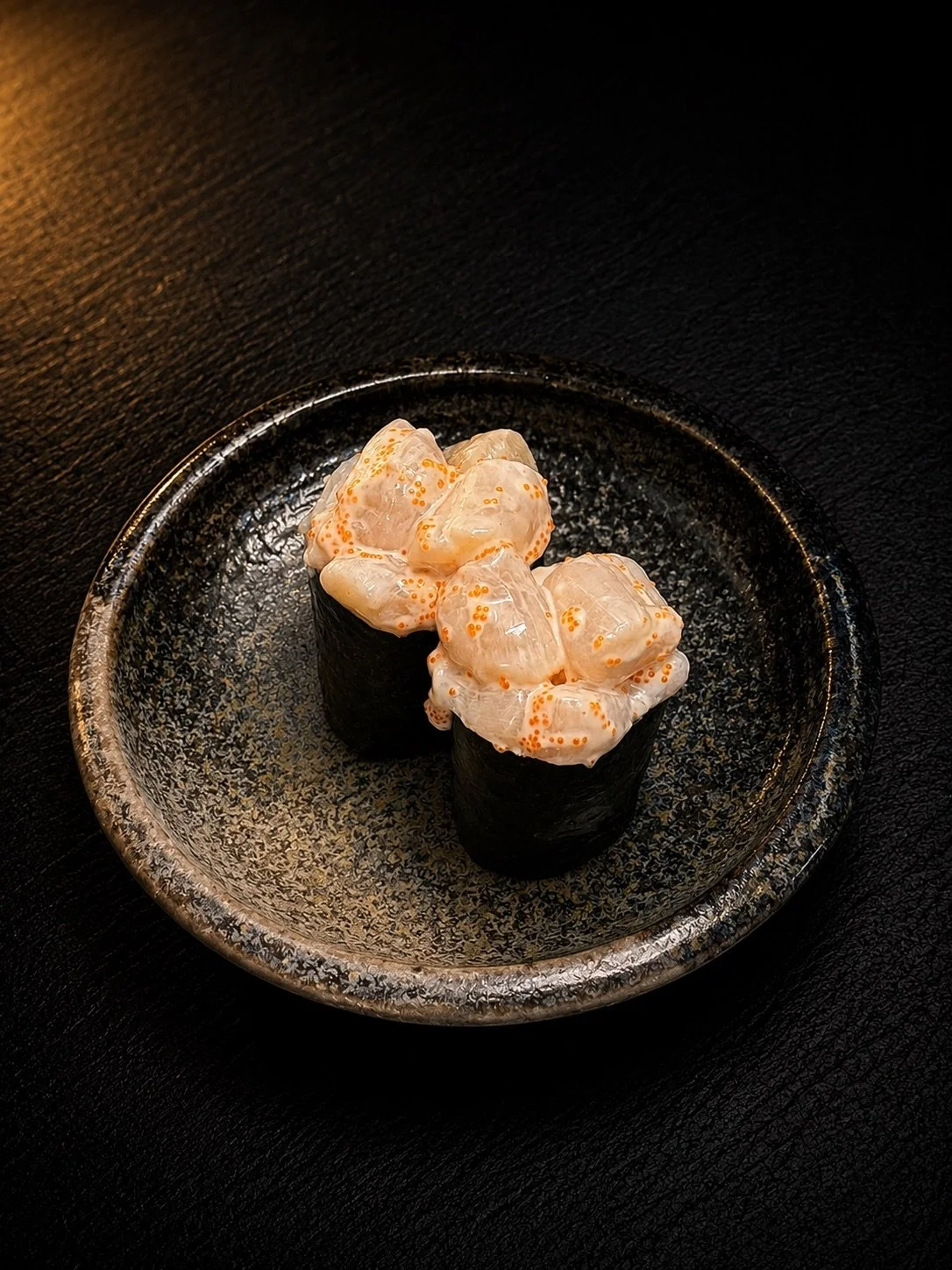 Two sushi rolls topped with spicy mayo and orange roe, on a black textured plate.