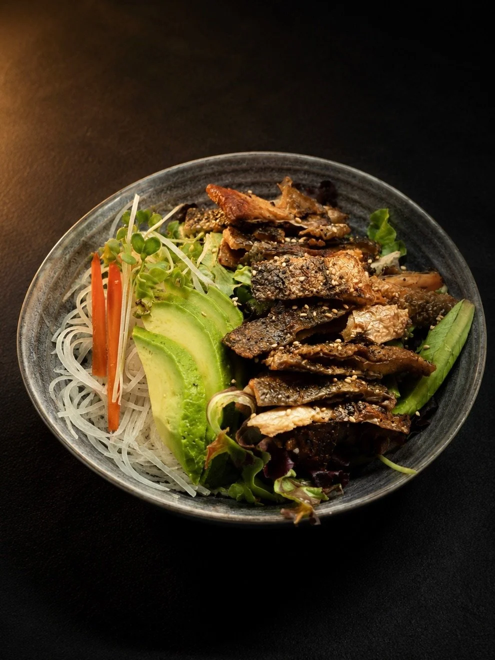 Bowl of salad with grilled fish, avocado slices, shredded carrots, bean sprouts, and mixed greens on a dark background.