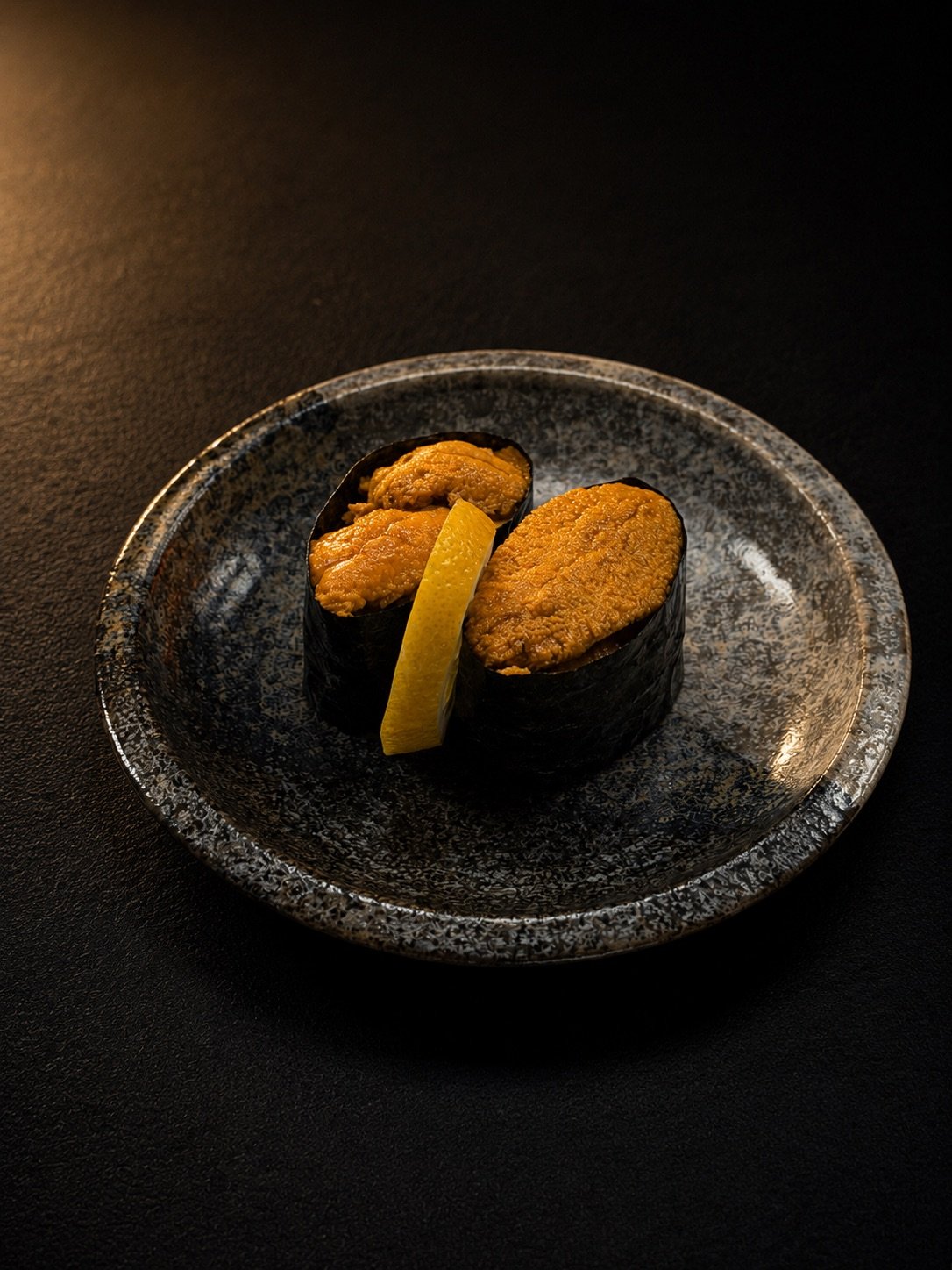 Two pieces of sushi topped with sea urchin, garnished with a lemon wedge, on a black ceramic plate.