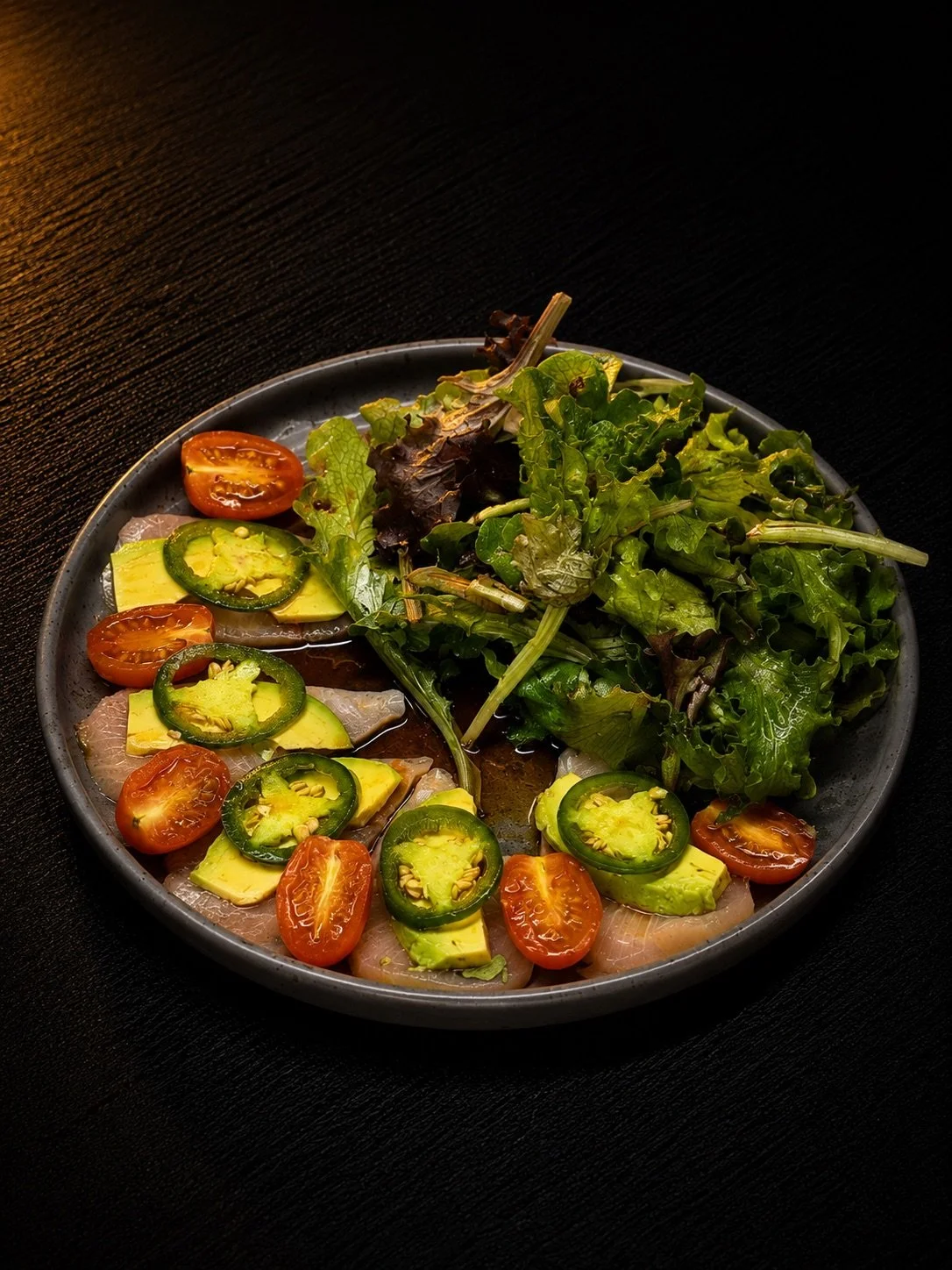 A plate of fresh salad with cherry tomatoes, avocado slices, cucumber slices, and mixed greens on a dark surface.