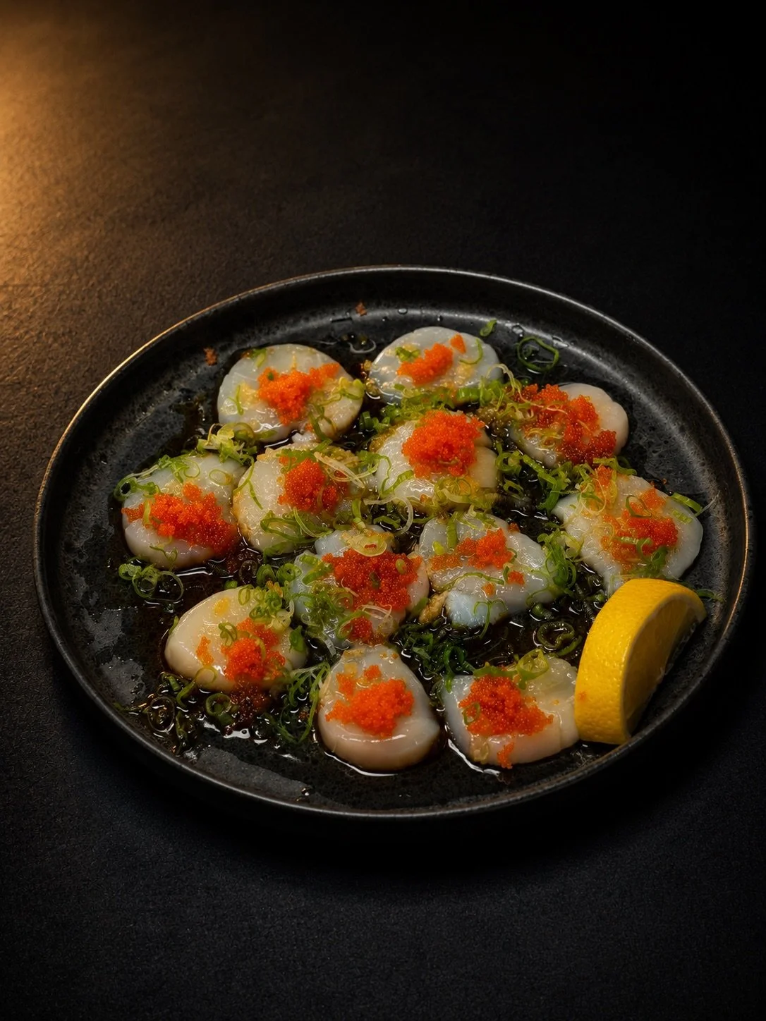 Plate of scallops topped with green onions, orange fish roe, and lemon wedge on black background.