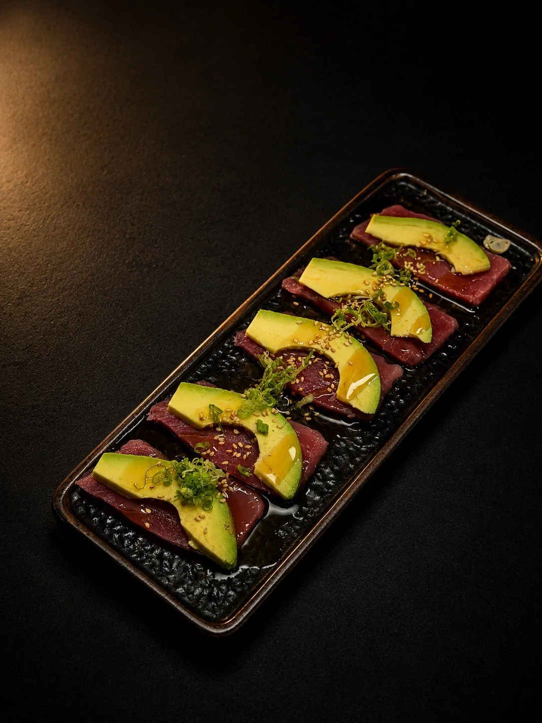 Sliced raw beef topped with sliced avocado, green onions, sesame seeds, and drizzle of sauce on a black rectangular plate.