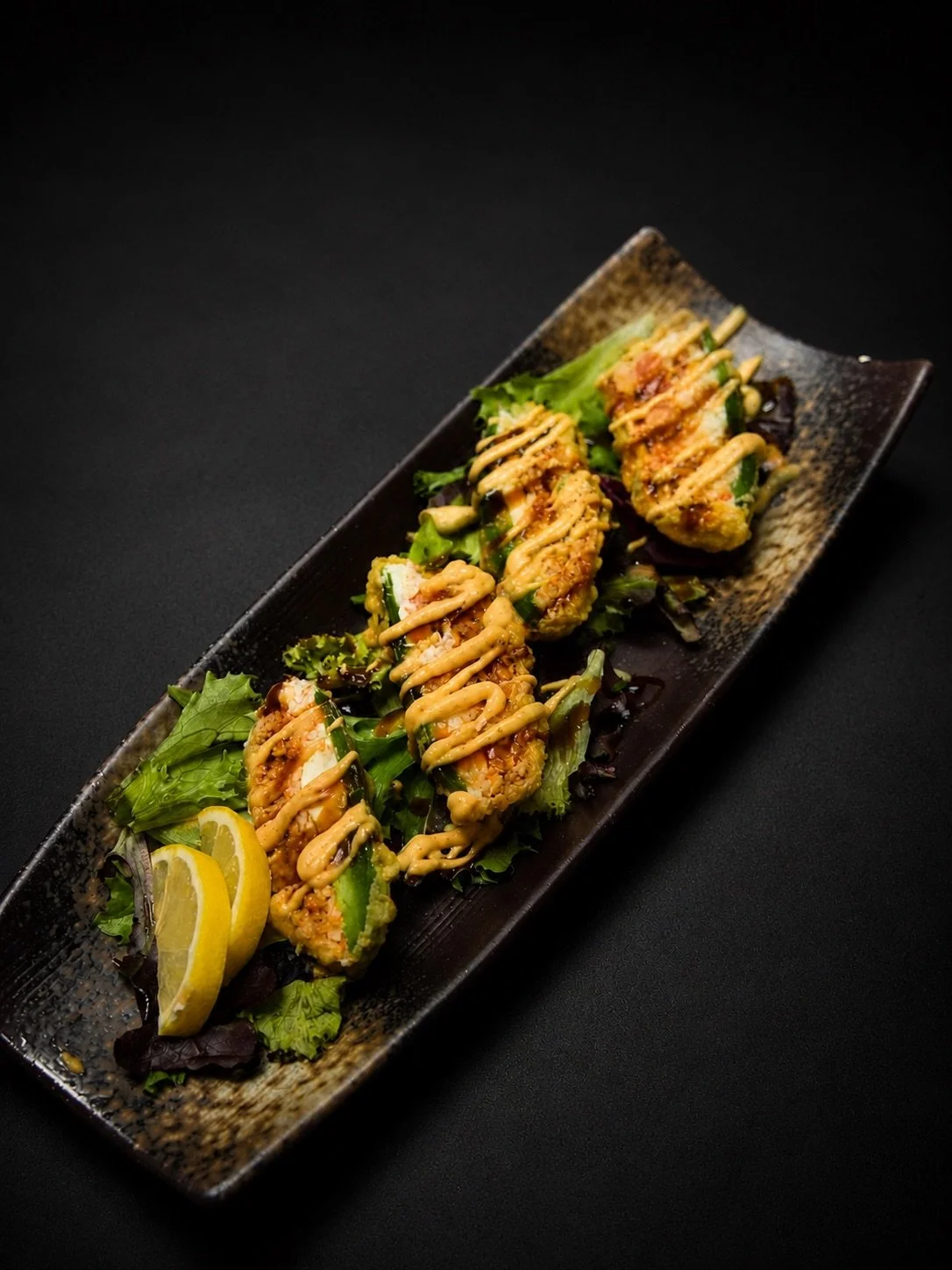 Sushi rolls topped with spicy mayo sauce, served on a black rectangular plate with leafy greens and lemon wedges, against a black background.