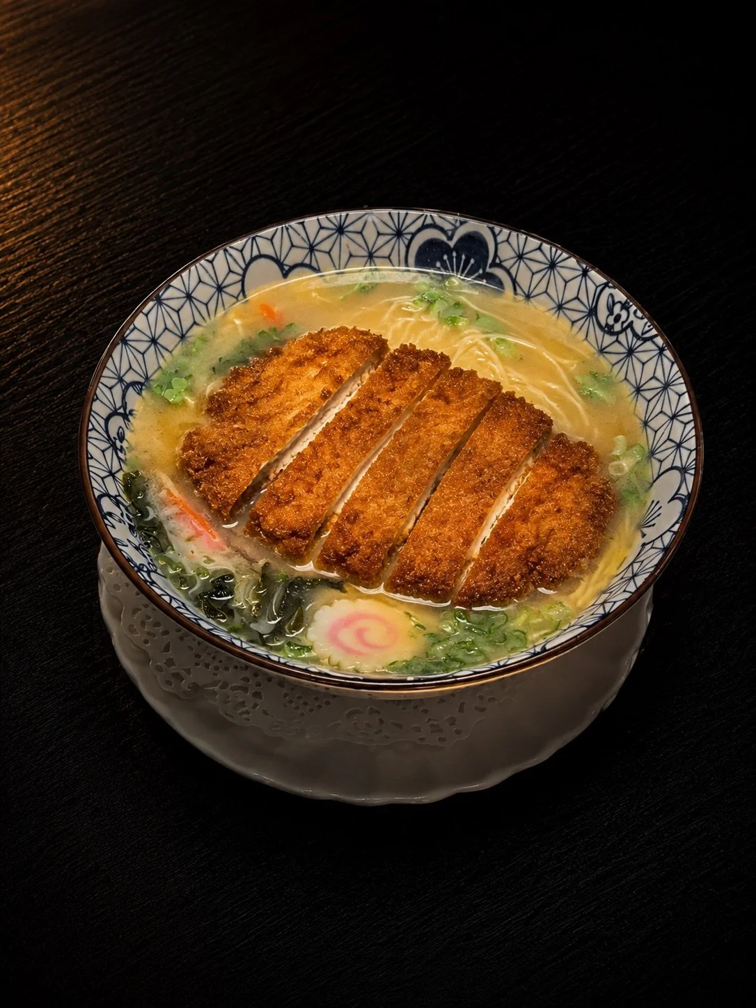A bowl of ramen with breaded fried cutlet on top, garnished with green onions and a narutomaki fish cake, in a patterned bowl on a dark table.