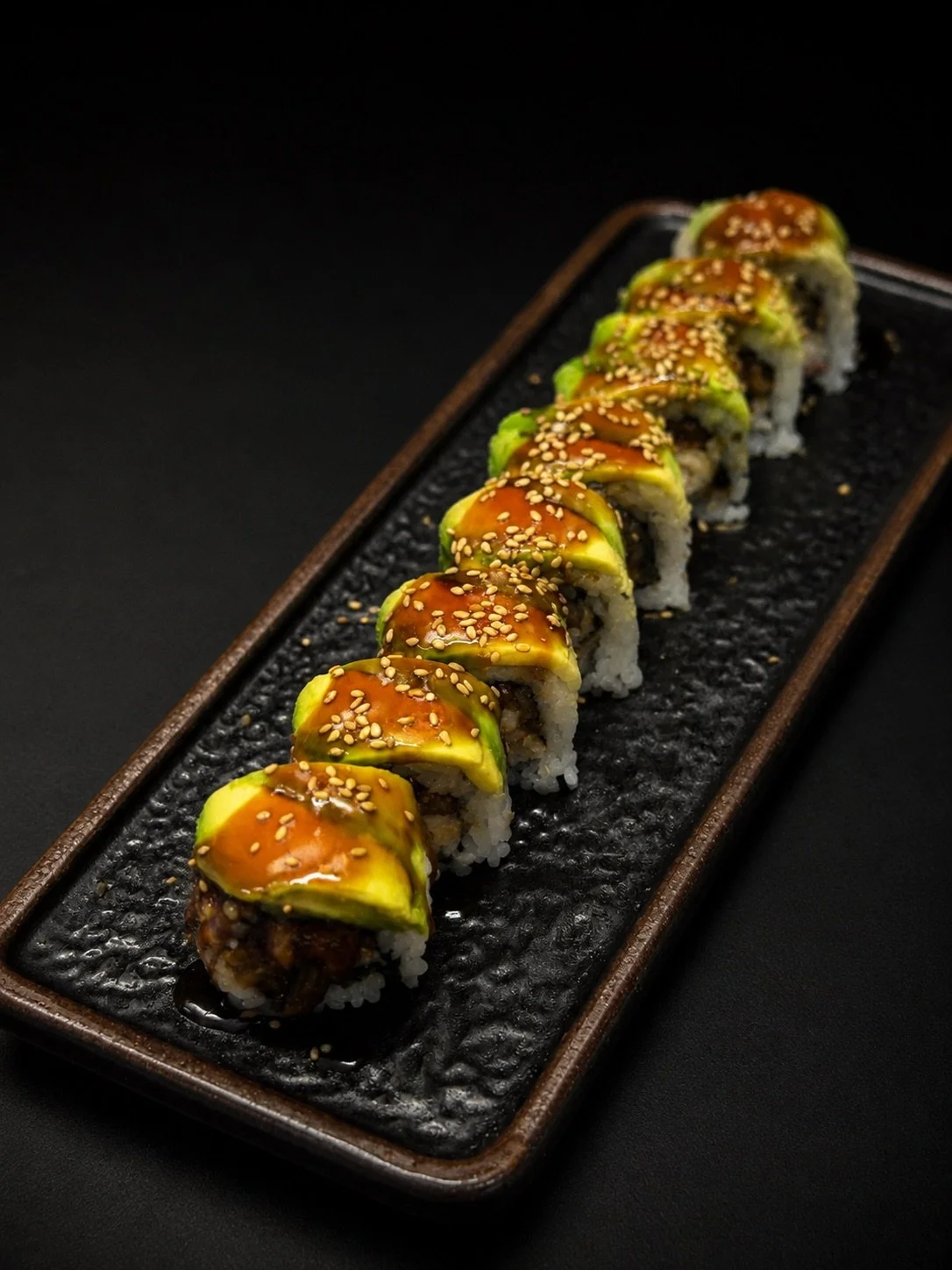 A row of sushi rolls topped with avocado slices, sesame seeds, and drizzled with sauce on a black rectangular plate.