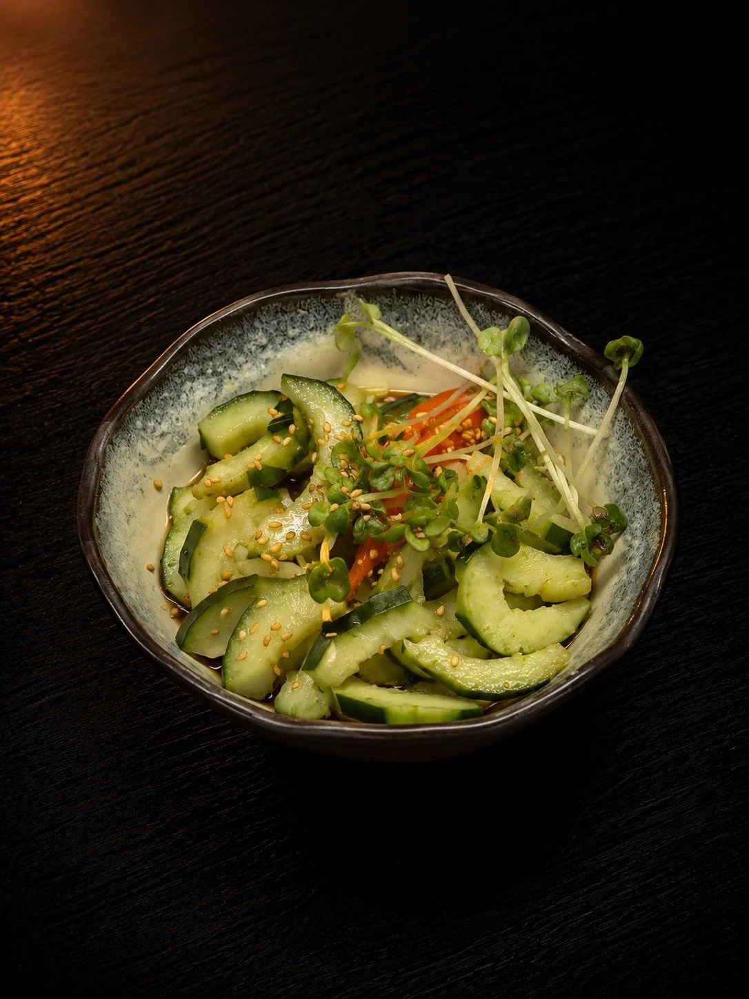 A small bowl of Asian cucumber salad with sesame seeds and microgreens on a black wooden surface.