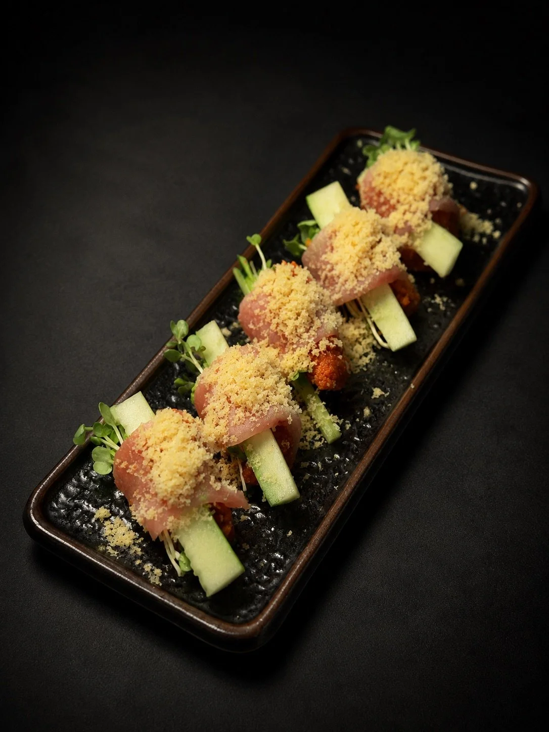 A rectangular black plate with five pieces of appetizer, each wrapped in thin slices of pink meat, topped with yellow crumbs, served over cucumber sticks and garnished with microgreens.