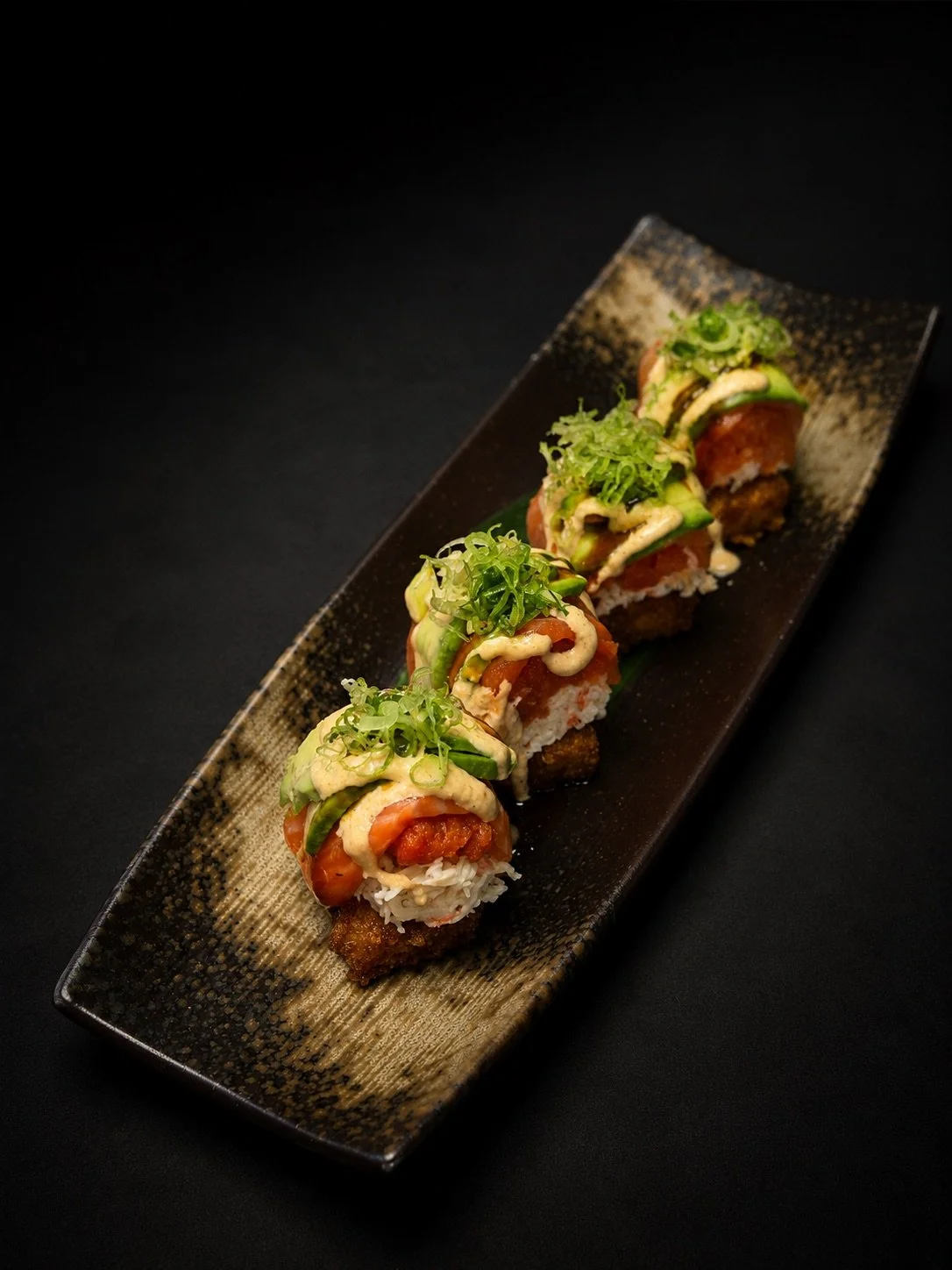 A plate of sushi rolls topped with avocado, green onions, and sauce, arranged on a black rectangular plate.