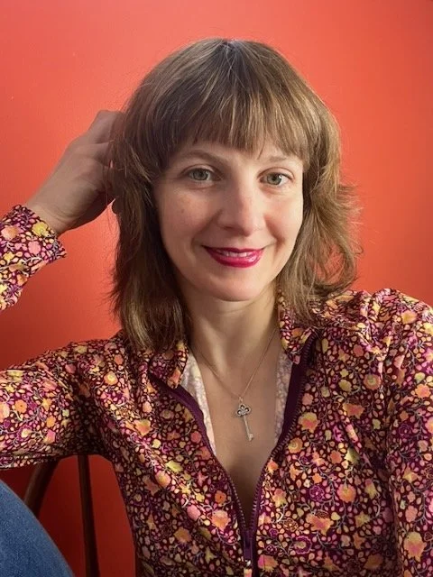 A woman with shoulder-length light brown hair wearing a patterned jacket with pink, purple, and yellow colors, smiling with red lipstick, in front of an orange background.