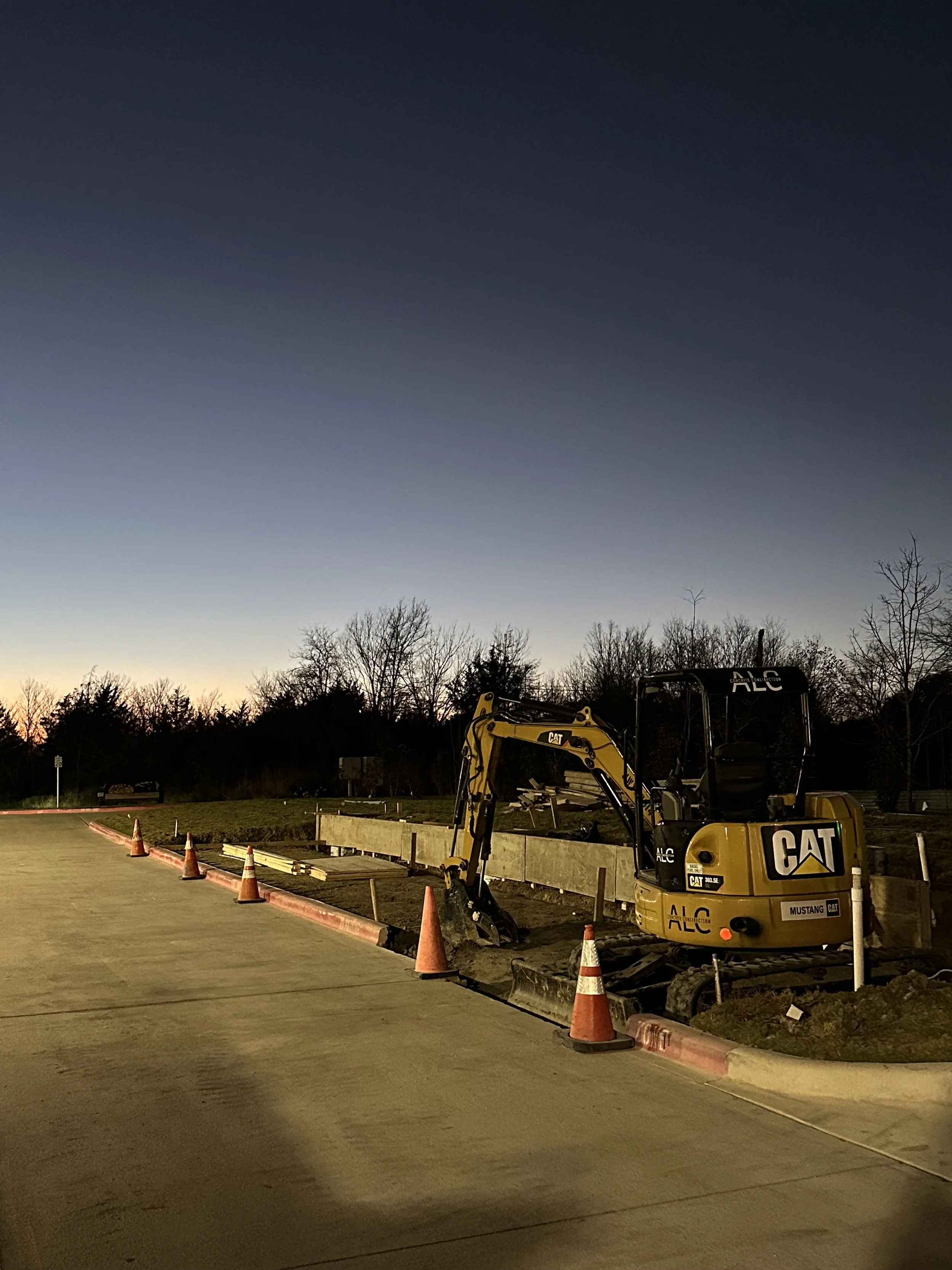 Construction site with a small yellow Caterpillar excavator surrounded by orange traffic cones and orange barriers, with trees and a darkening sky in the background.