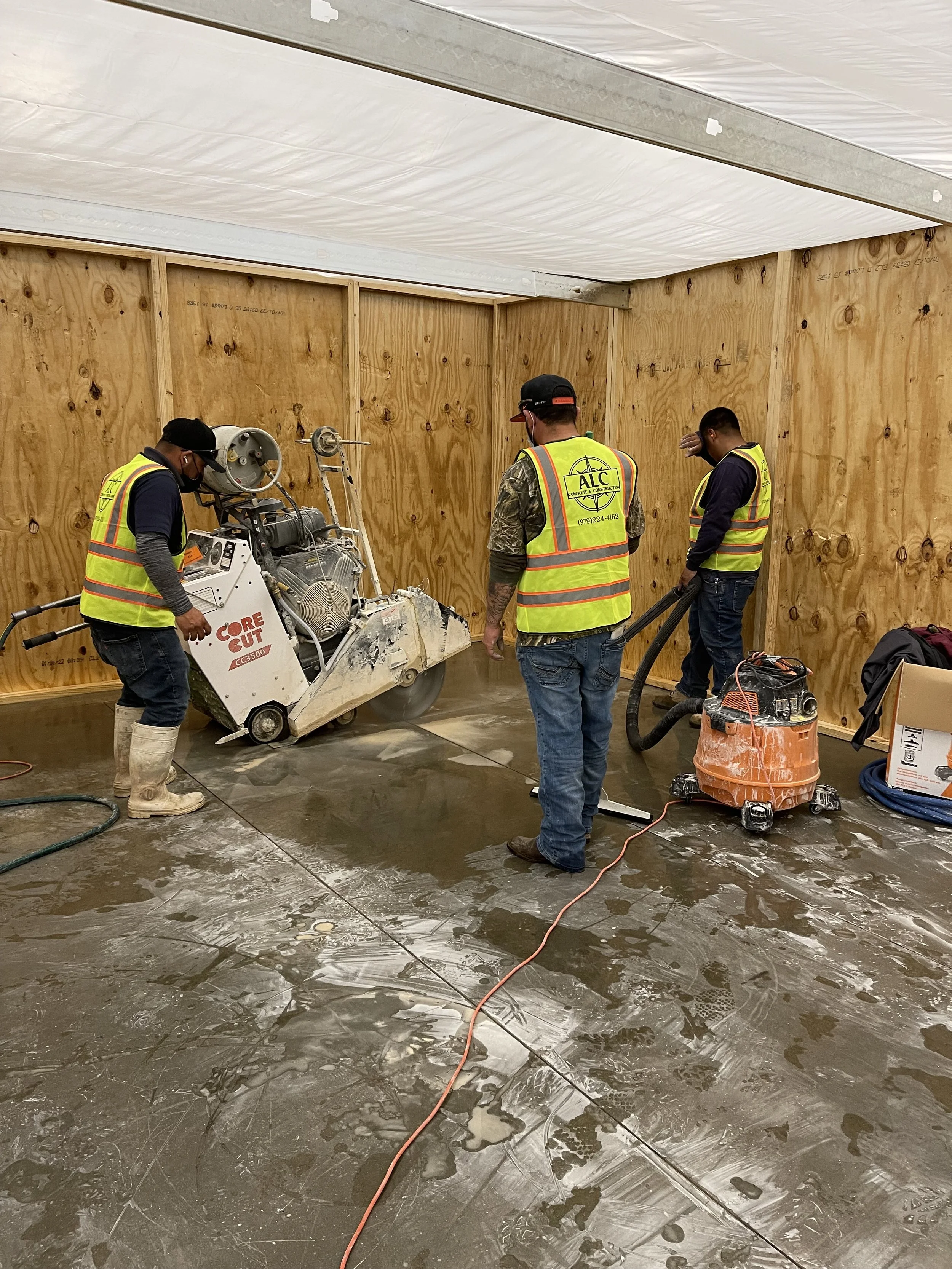 Workers in high-visibility vests working on a concrete floor with a large commercial retail store l, surrounded by construction tools and equipment inside a room under construction with plywood walls and a ceiling covered in plastic sheeting.