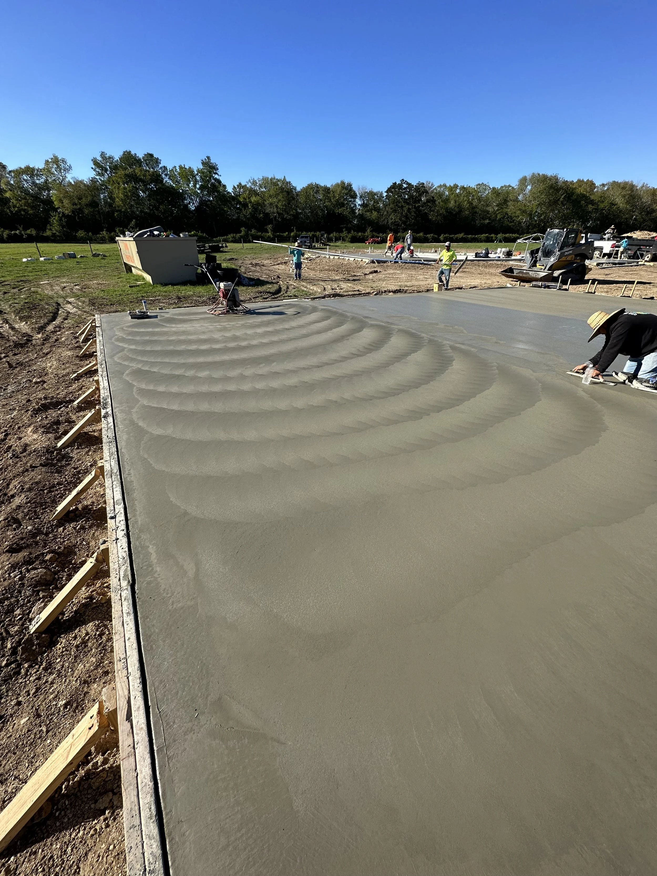 Workers pouring and smoothing concrete on a construction site with construction equipment and trees in the background.