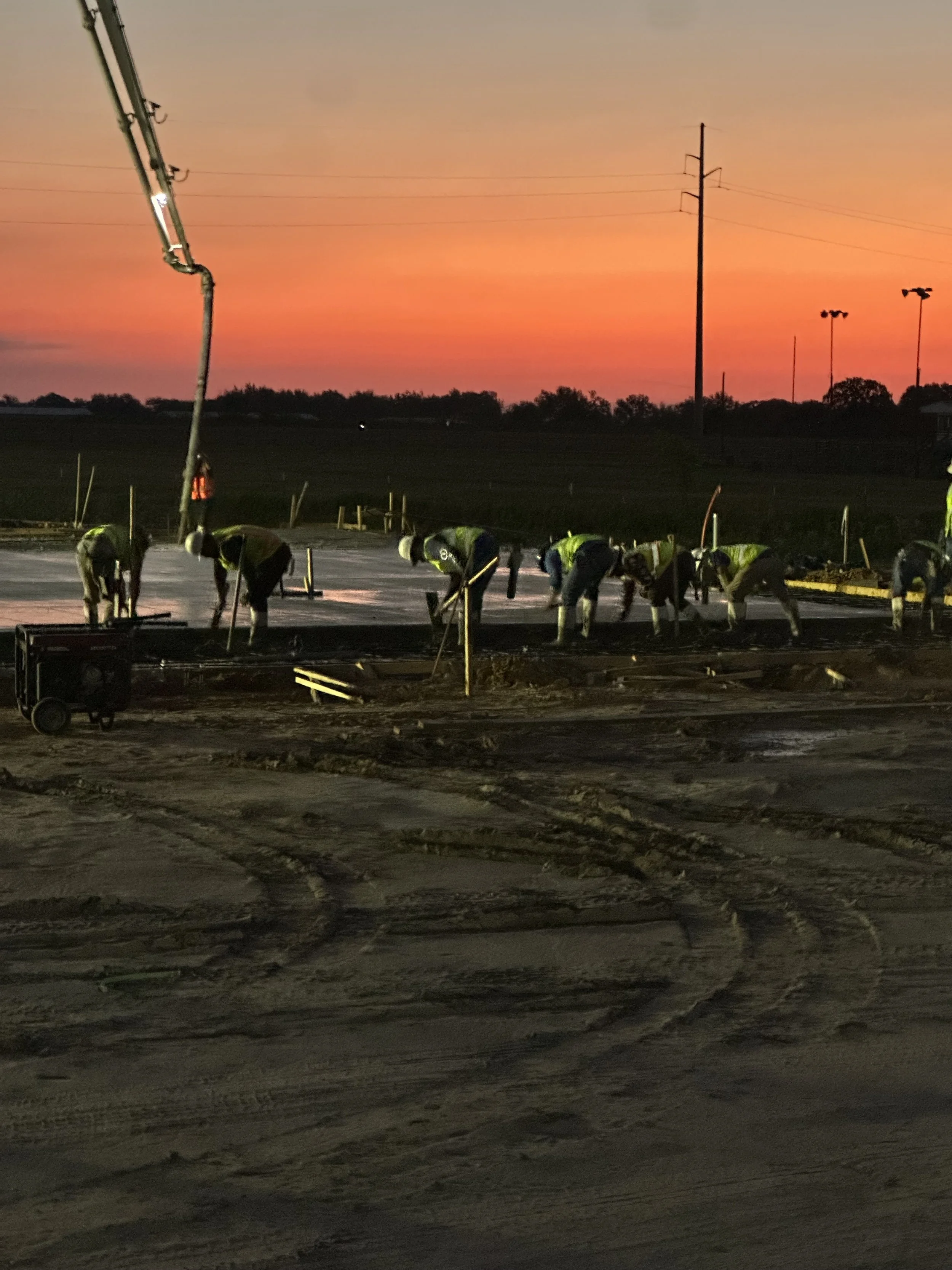 Construction workers laying down fresh concrete on a road at sunset, with a hydraulic pump and power lines visible in the background.