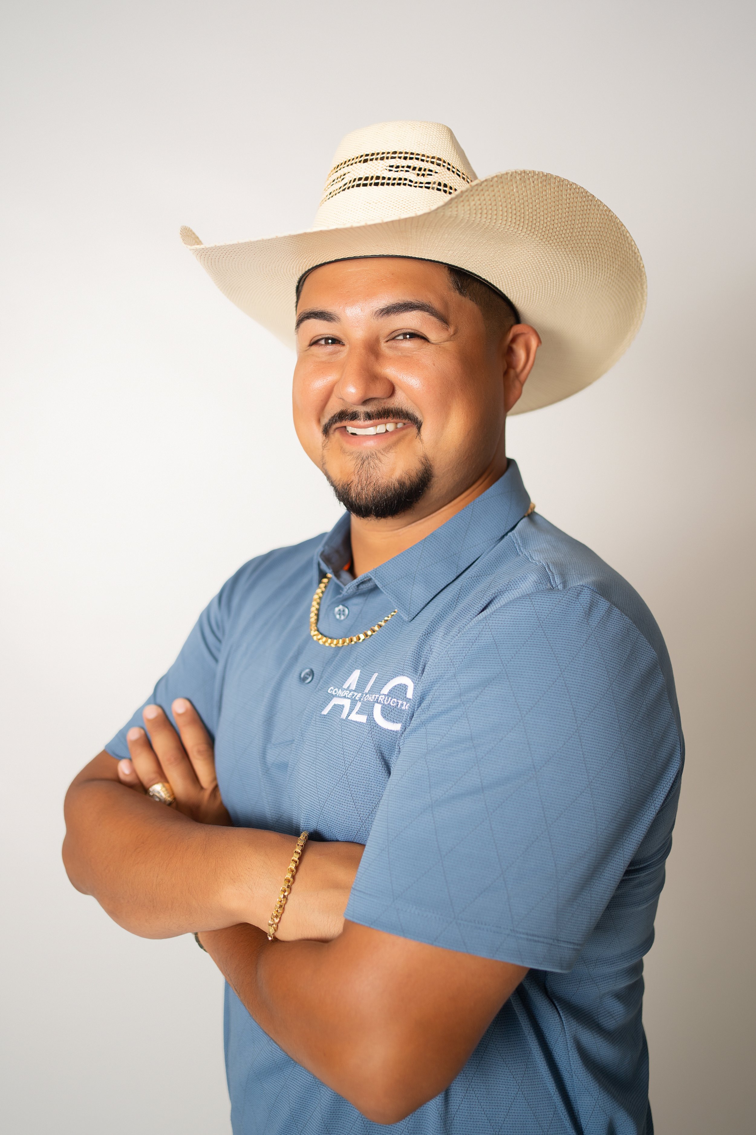 A man with a goatee and mustache, smiling and squinting slightly, wearing a wide-brimmed straw hat, a blue collared shirt, a gold chain necklace, and a gold bracelet, standing with crossed arms against a plain white background.