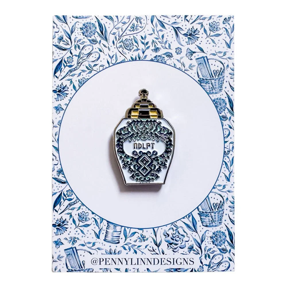 A decorative pin shaped like a perfume bottle, with intricate black and silver design and the word "IDLFT" in the center. The pin is mounted on a card with a blue floral pattern and the handle "@PENNYLINNDESIGNS" written at the bottom.