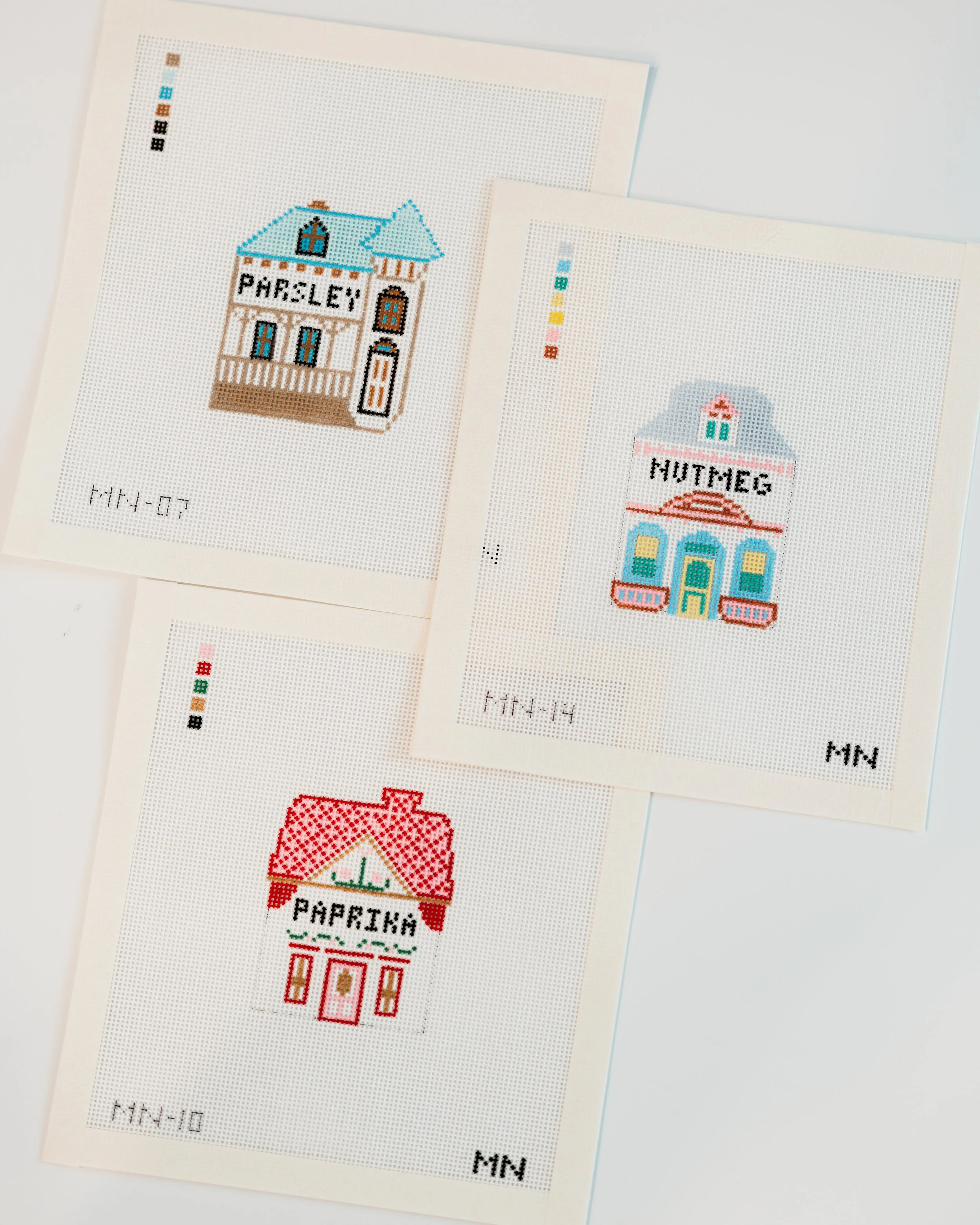 Two pieces of white paper with cross-stitch designs of houses, each labeled with the words 'PAPRIKA' and 'NUTMEG' respectively, on a white surface.