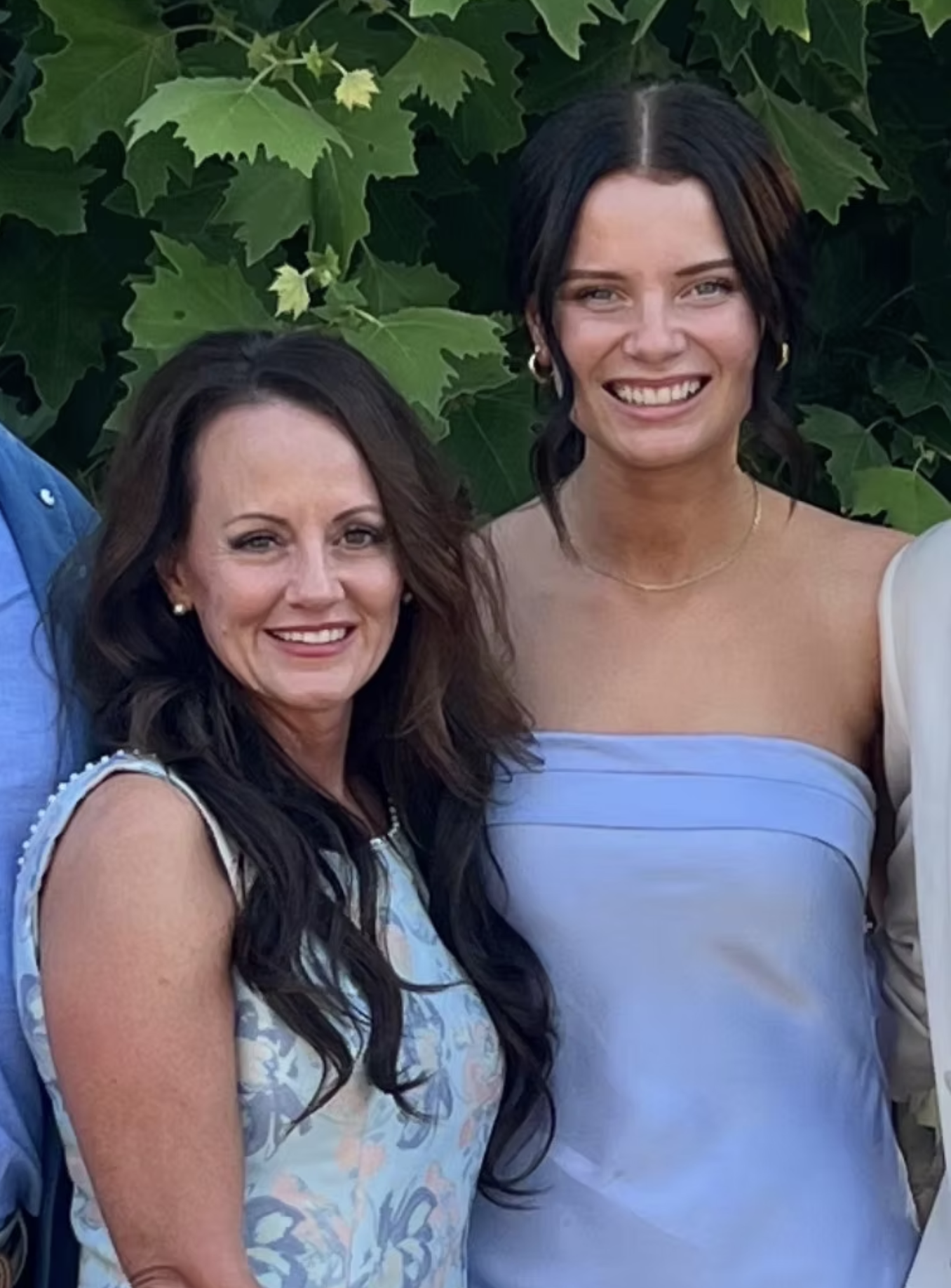 Smiling women in dresses posing outdoors with green leafy background.