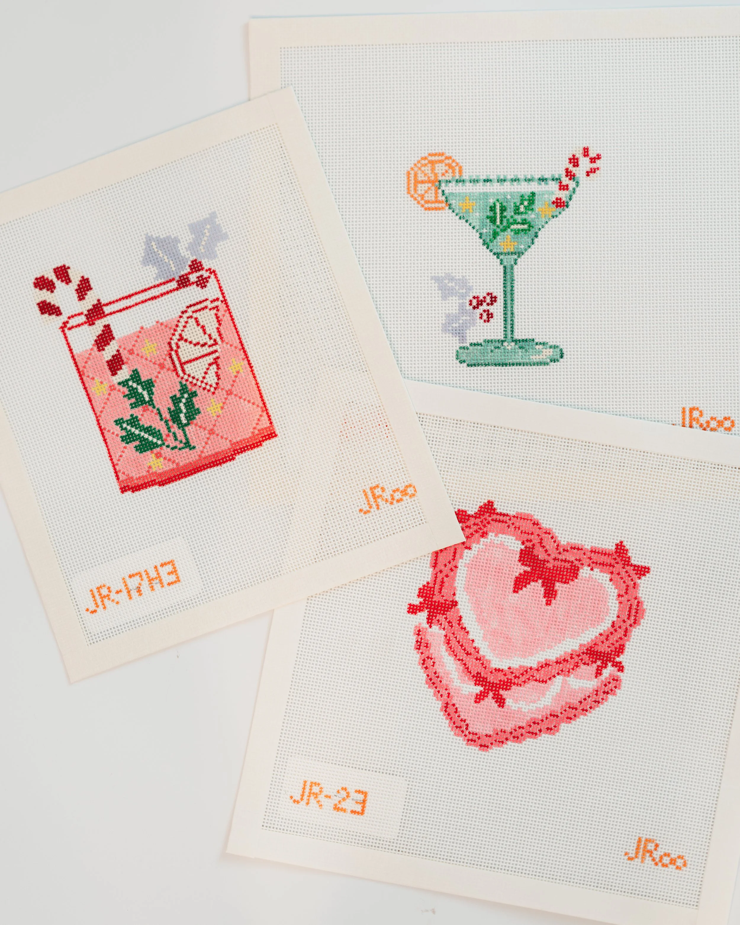 Two pieces of cross-stitch embroidery on white fabric, featuring colorful images of a cocktail glass with a slice of lemon and a cherry, and a red heart with a pearl necklace design, marked with orange thread labels 'Jr-23' and 'Jr-24'.