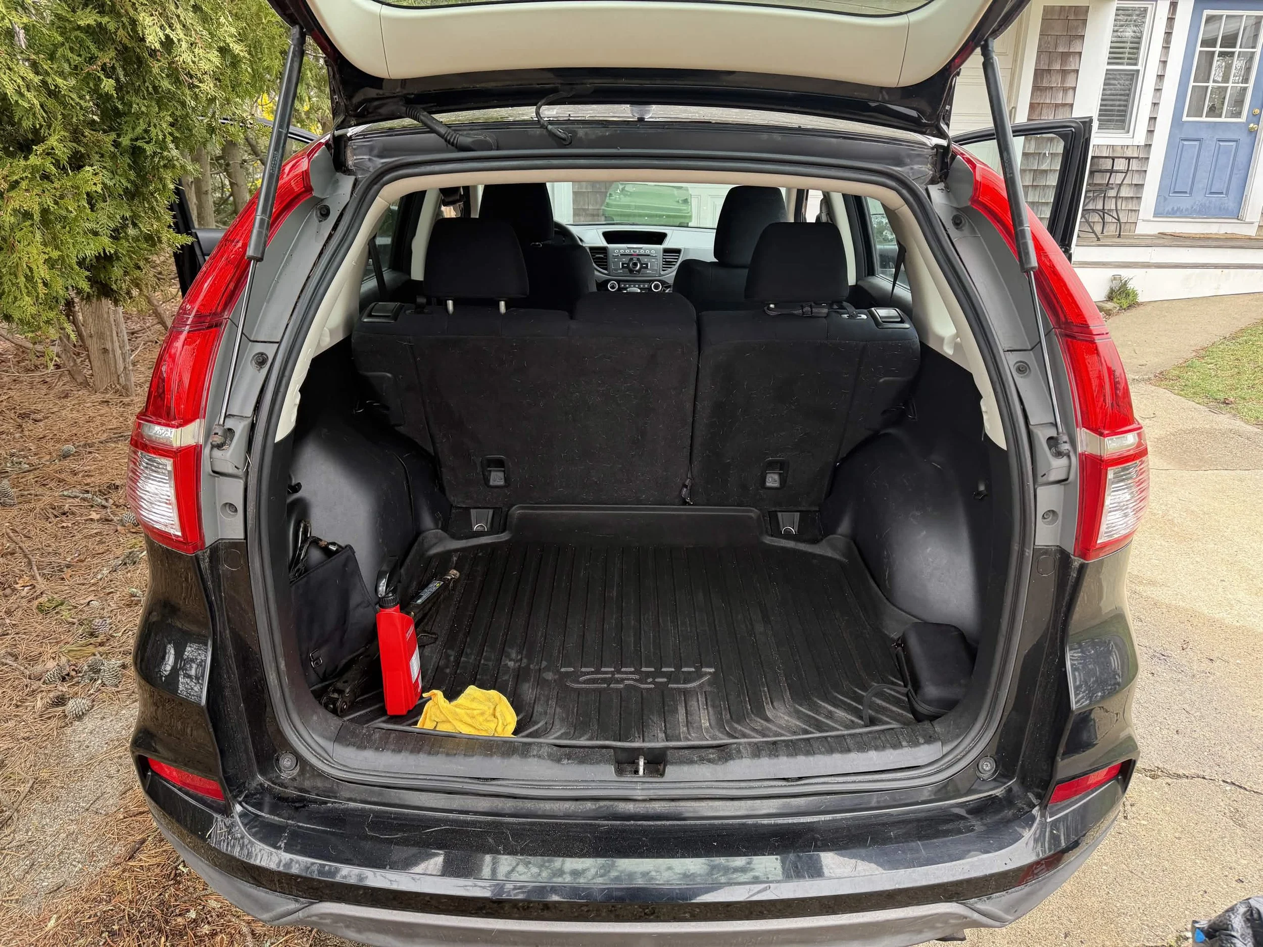 The open trunk of a black SUV parked outside near a house, showing the rear interior with a rubber cargo mat, a red item, and a yellow cloth.