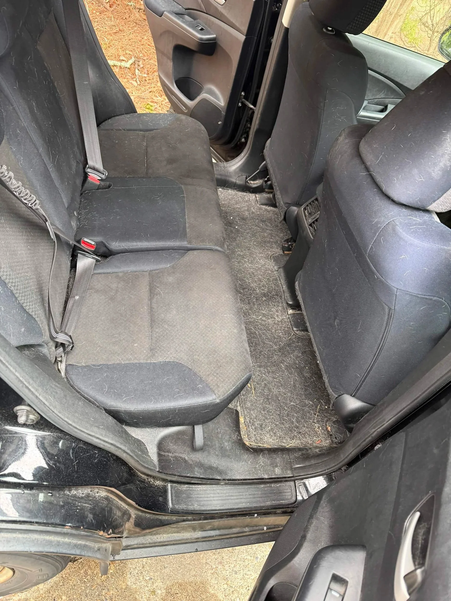 The backseat of a black vehicle with dusty and dirty black fabric seats and floor mats, showing signs of wear and dirt.