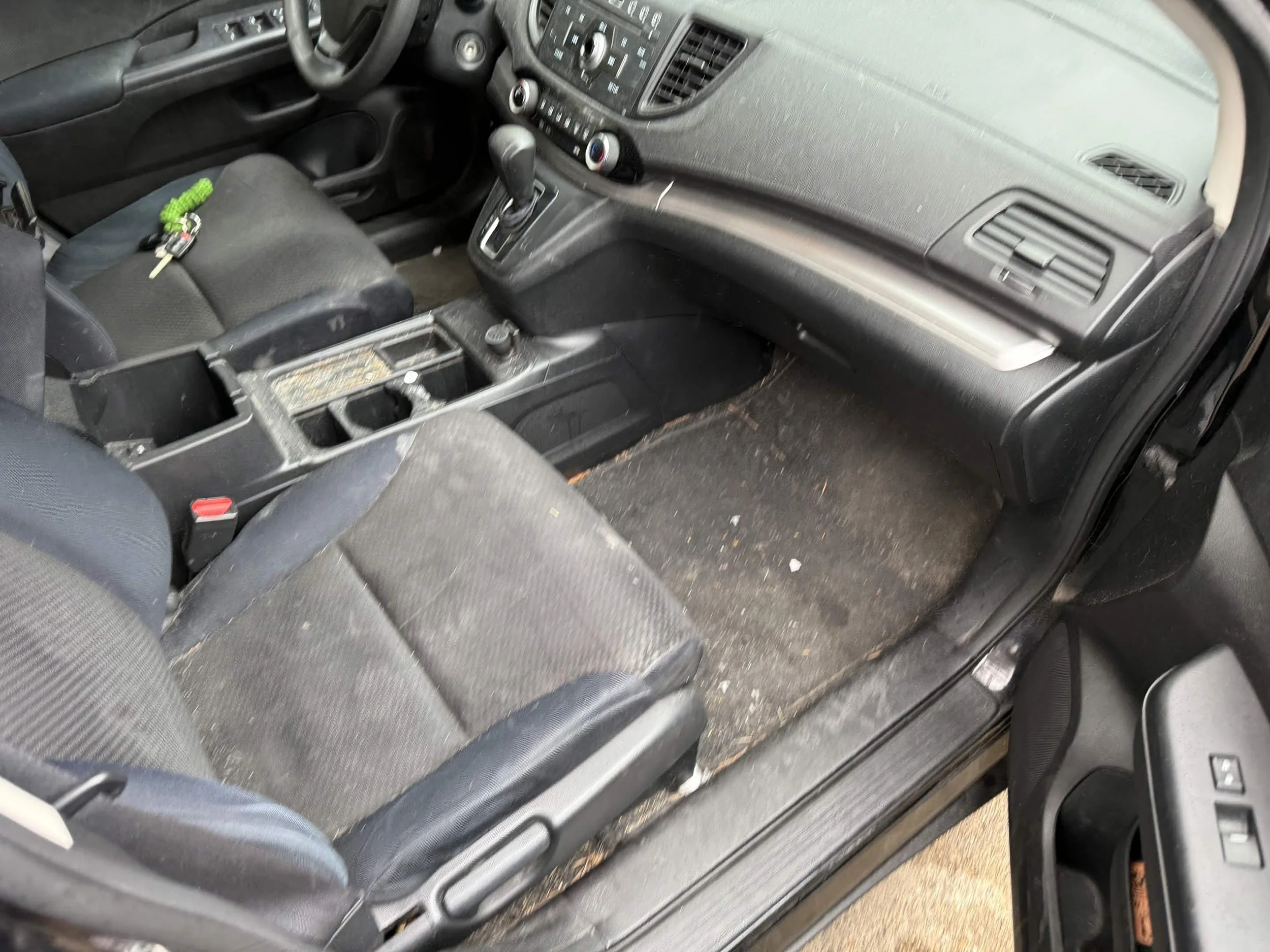 The interior of a used car showing the front passenger and driver seats, a center console with an open compartment, a gear shift, and a dusty floor with some dirt and debris.