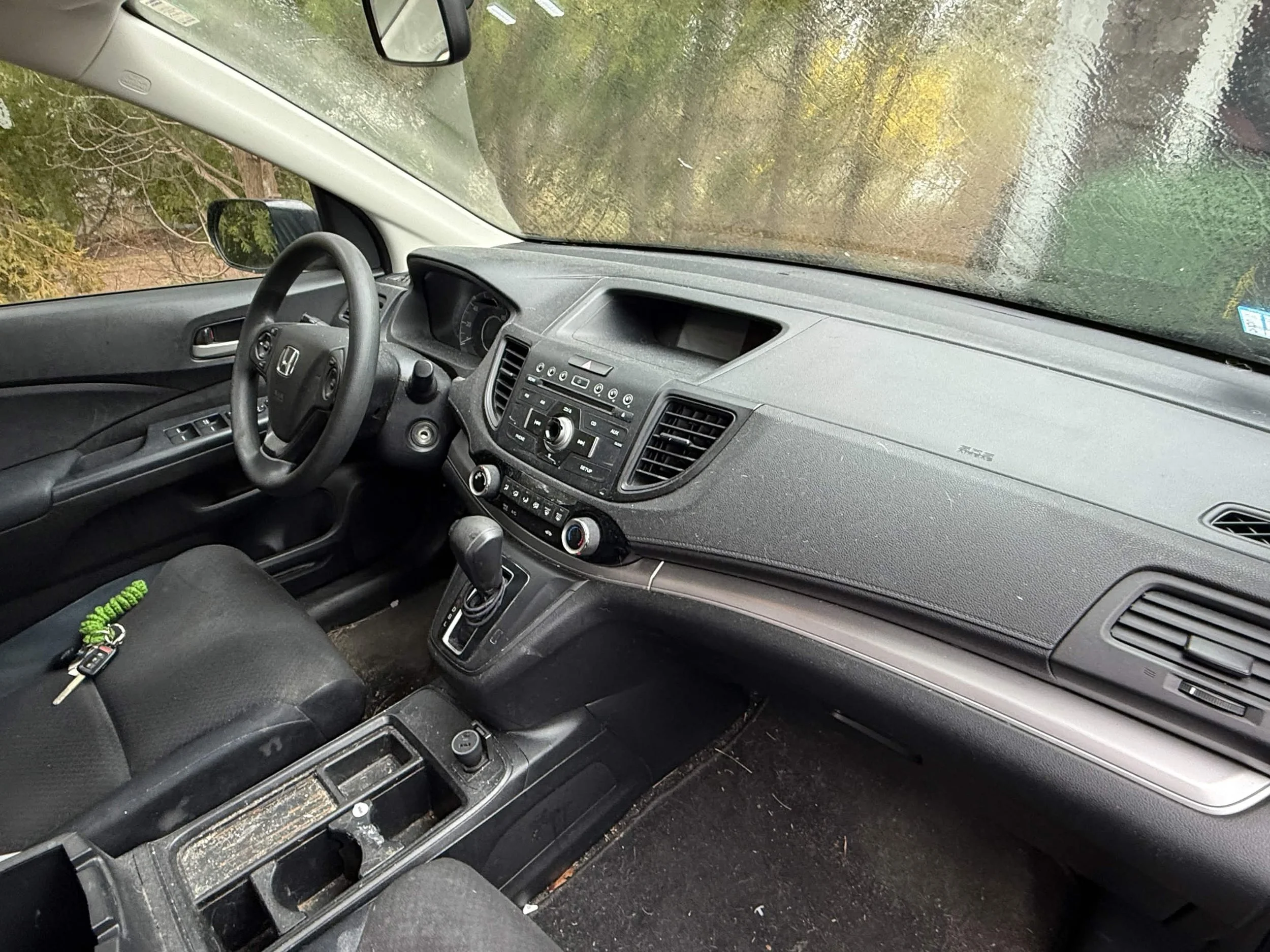 Inside view of a car's dashboard and front seats, showing the steering wheel, gear shift, and control panel with various buttons and dials, with a small keychain on the driver's seat.