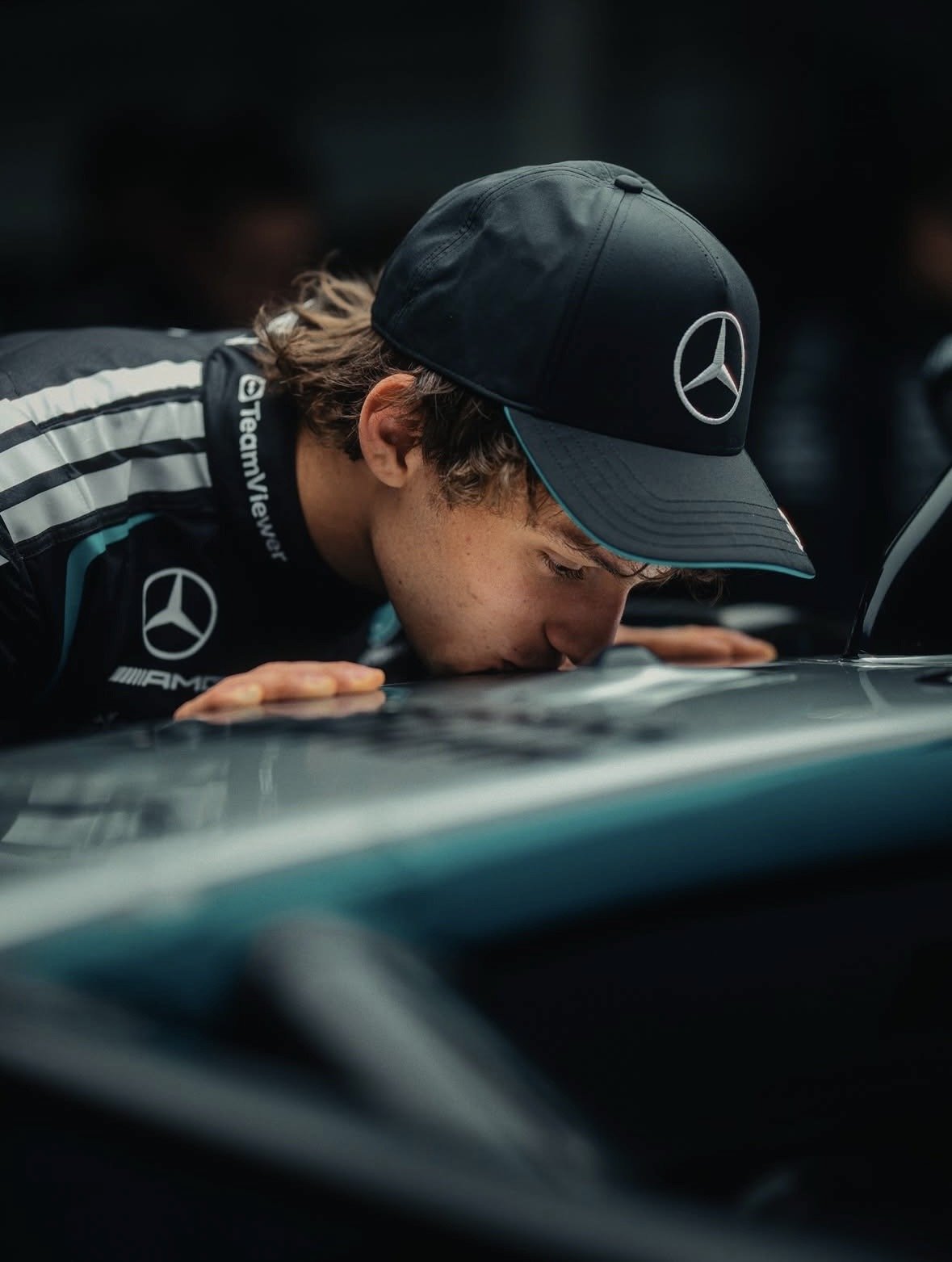 A young man in racing gear leans over the hood of a car, smelling or inspecting it. He wears a black cap with the Mercedes-Benz logo and a racing suit with Mercedes and AMG logos.