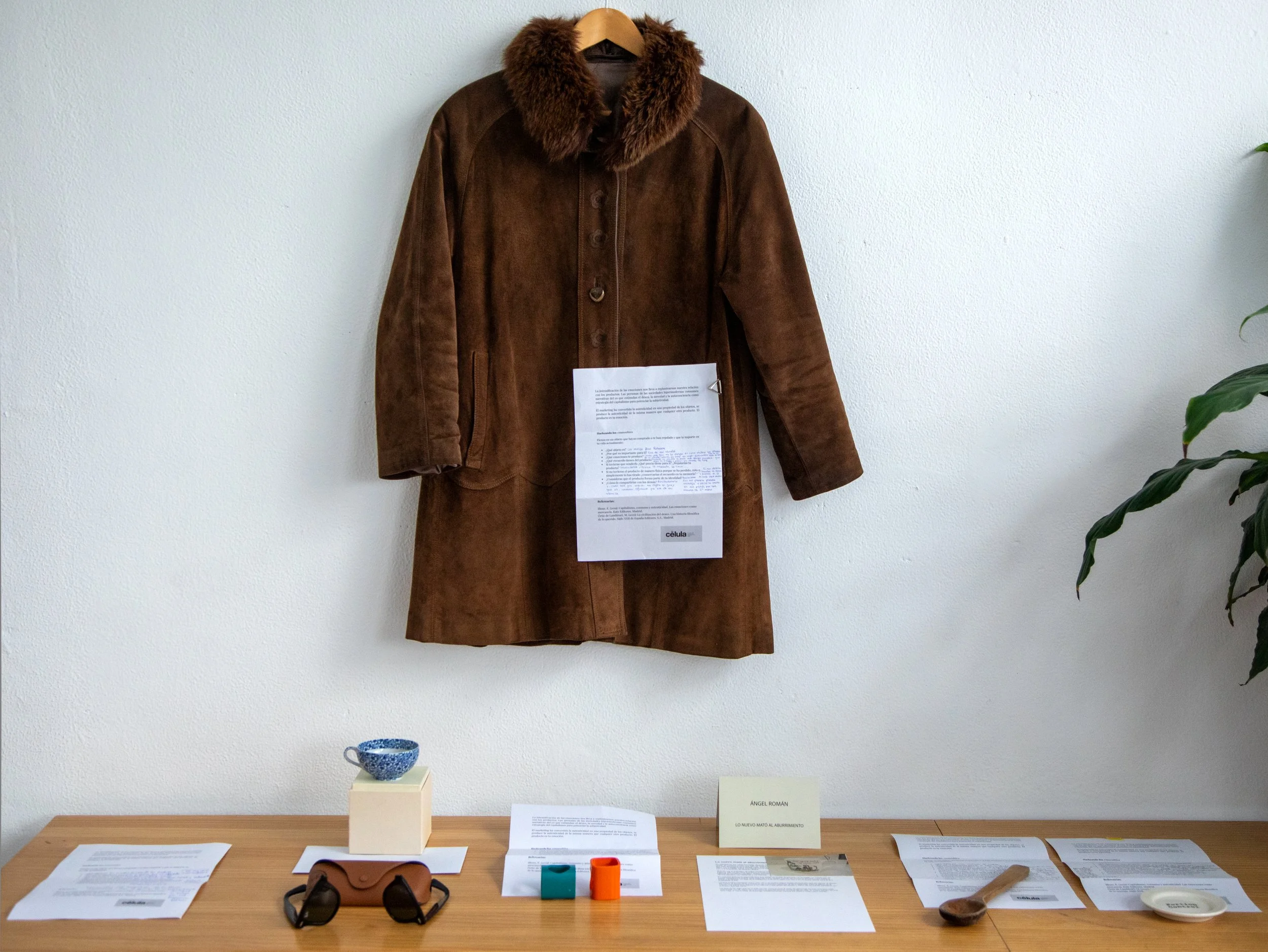 Exposición en un museo con una chamarra de piel marrón con cuello de piel, colgada en la pared. En la mesa hay varias hojas, documentos, un par de lentes, objetos de colores y cajas, con una planta a la derecha y un cartel que dice 'ÁNGEL ROMÁN'.