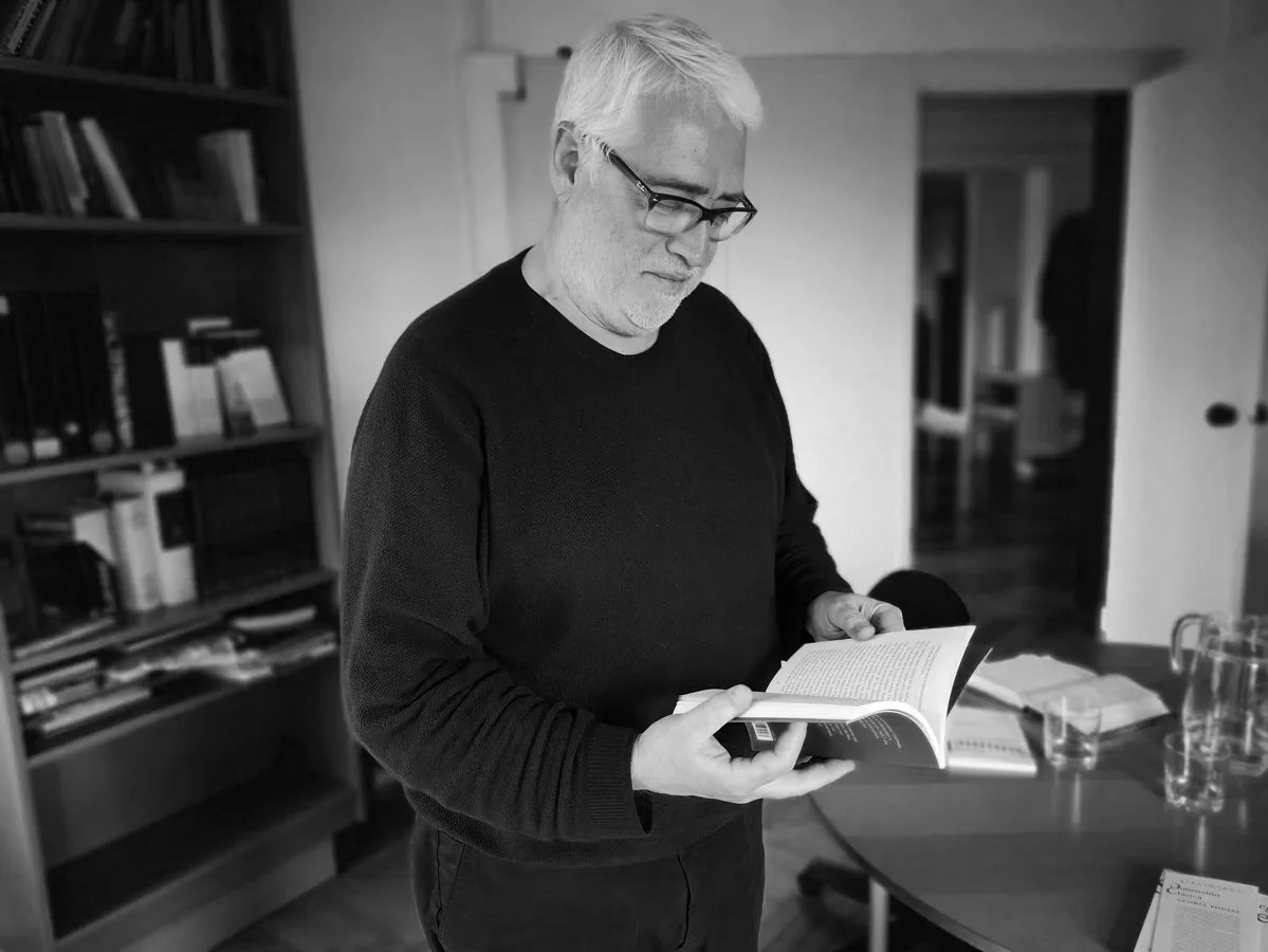 Hombre de cabello gris y gafas leyendo un libro en una oficina o salón de estudio, con estantería de libros y una mesa con papeles y vasos al fondo.