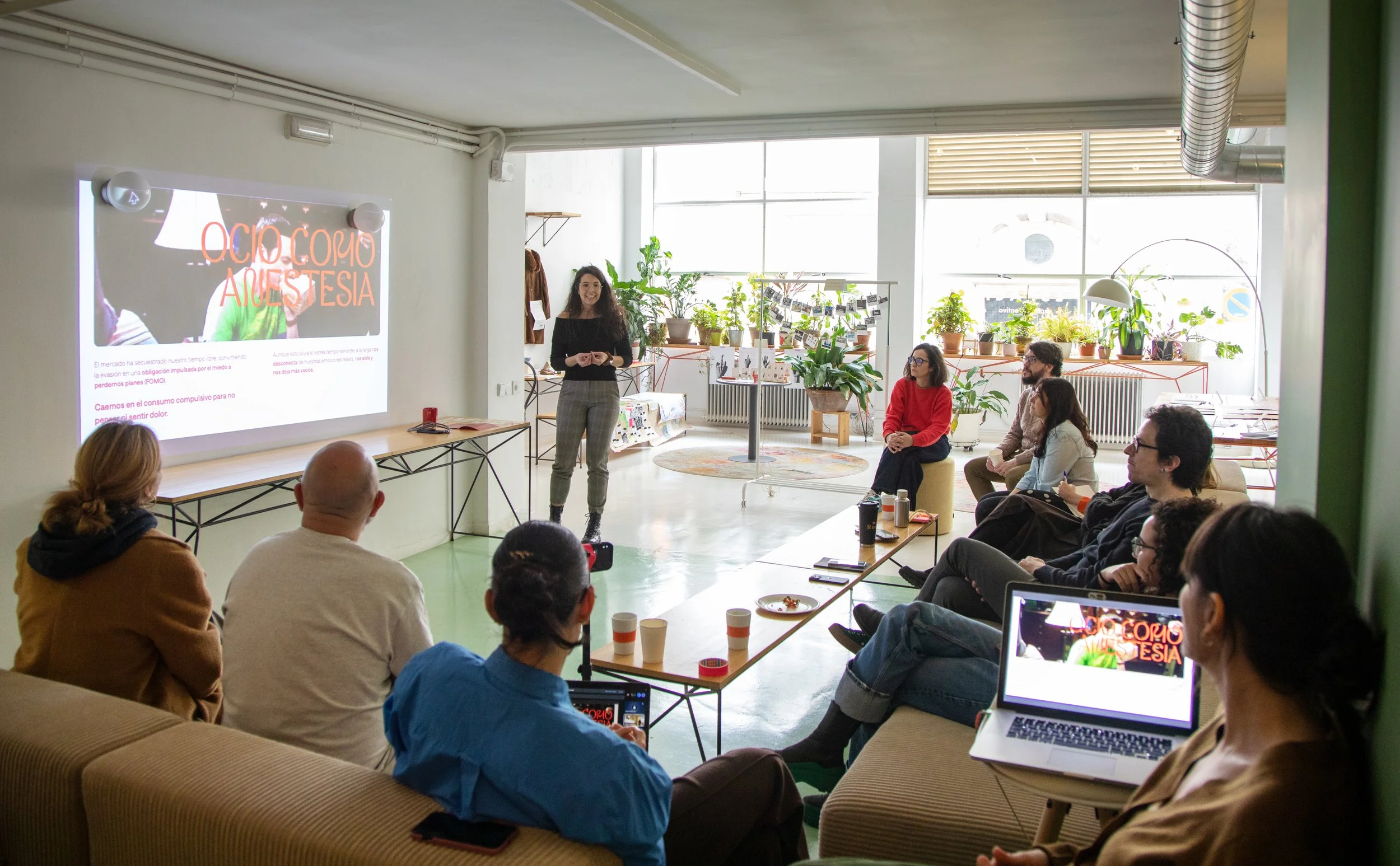 Una mujer da una presentación a un grupo de personas en un ambiente de oficina con muchas plantas y una pantalla grande que dice "Ojo como autestesia".