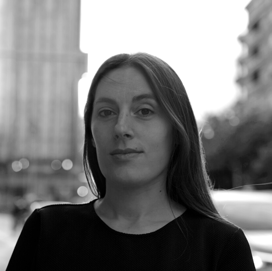 Retrato en blanco y negro de una mujer con cabello largo y oscuro, en un entorno urbano con edificios y árboles difusos de fondo.