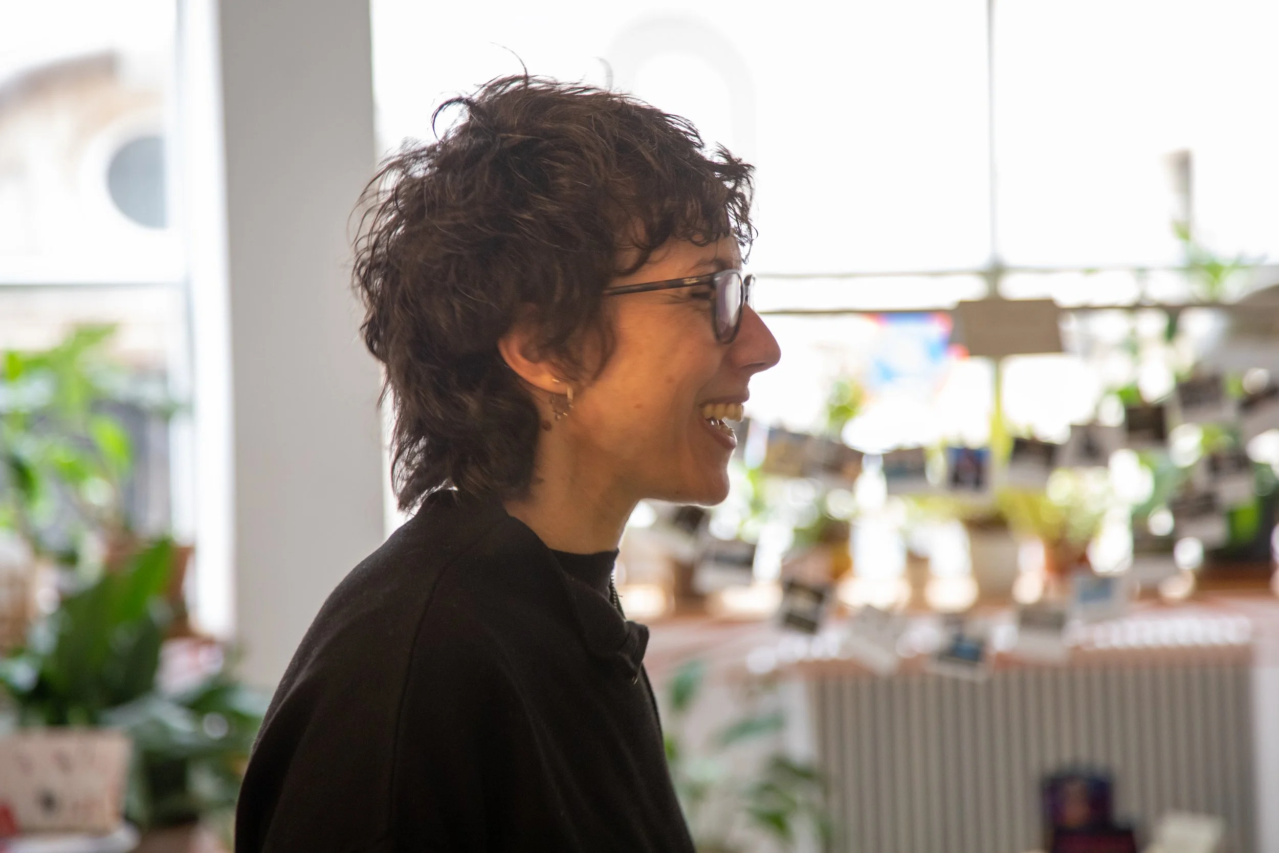 Mujer con gafas sonriendo, en un entorno interior con muchas plantas y luz natural.
