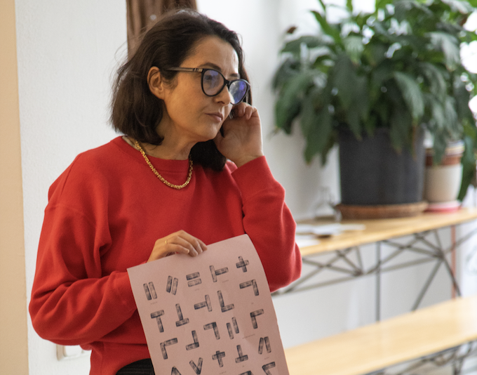 Mujer con gafas y sudadera roja sosteniendo una hoja con letras en un entorno interior con plantas y muebles de madera.