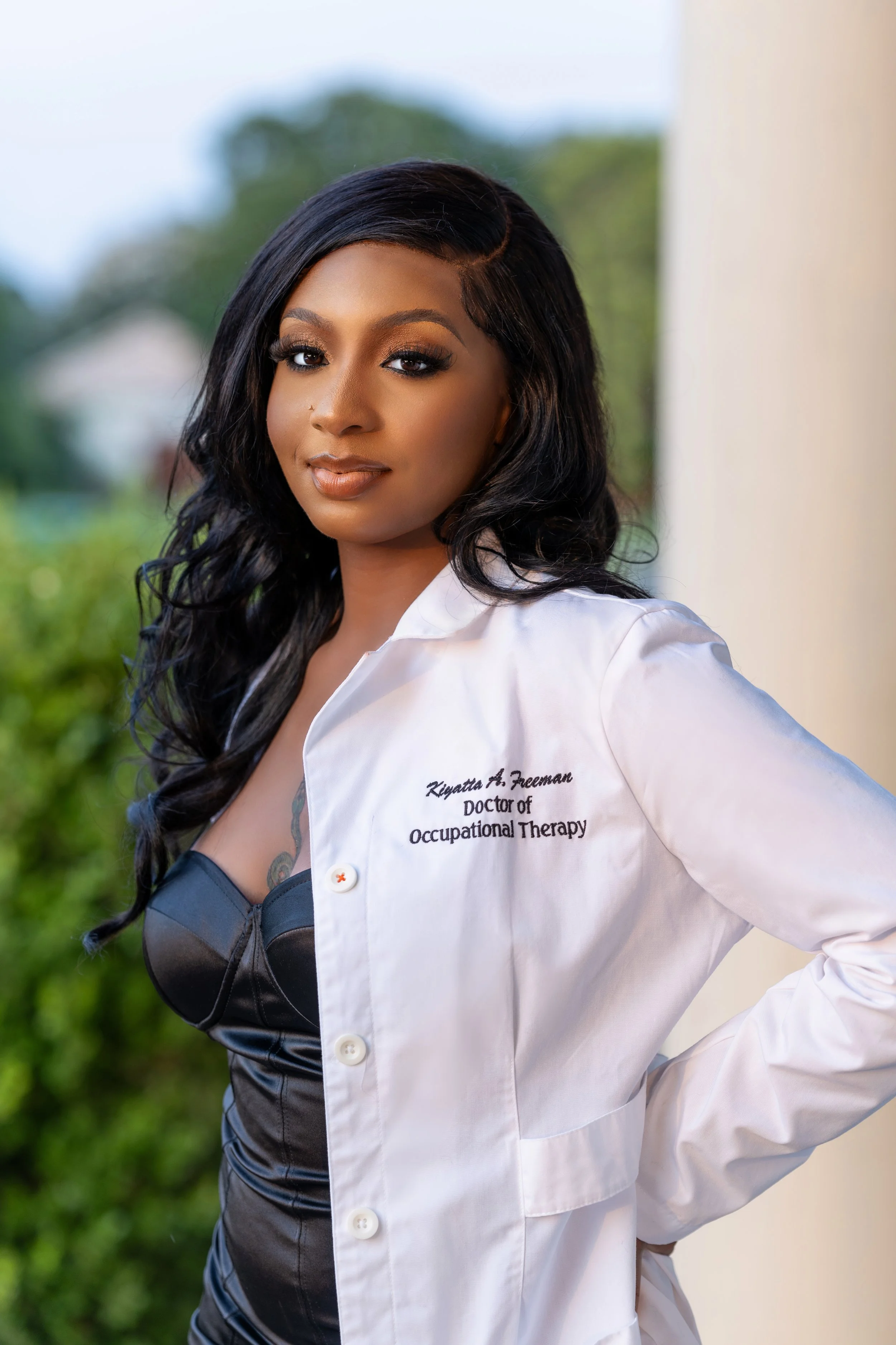 A woman with long black wavy hair wearing a white lab coat with embroidered text and a black dress underneath, standing outdoors with a blurred green background.