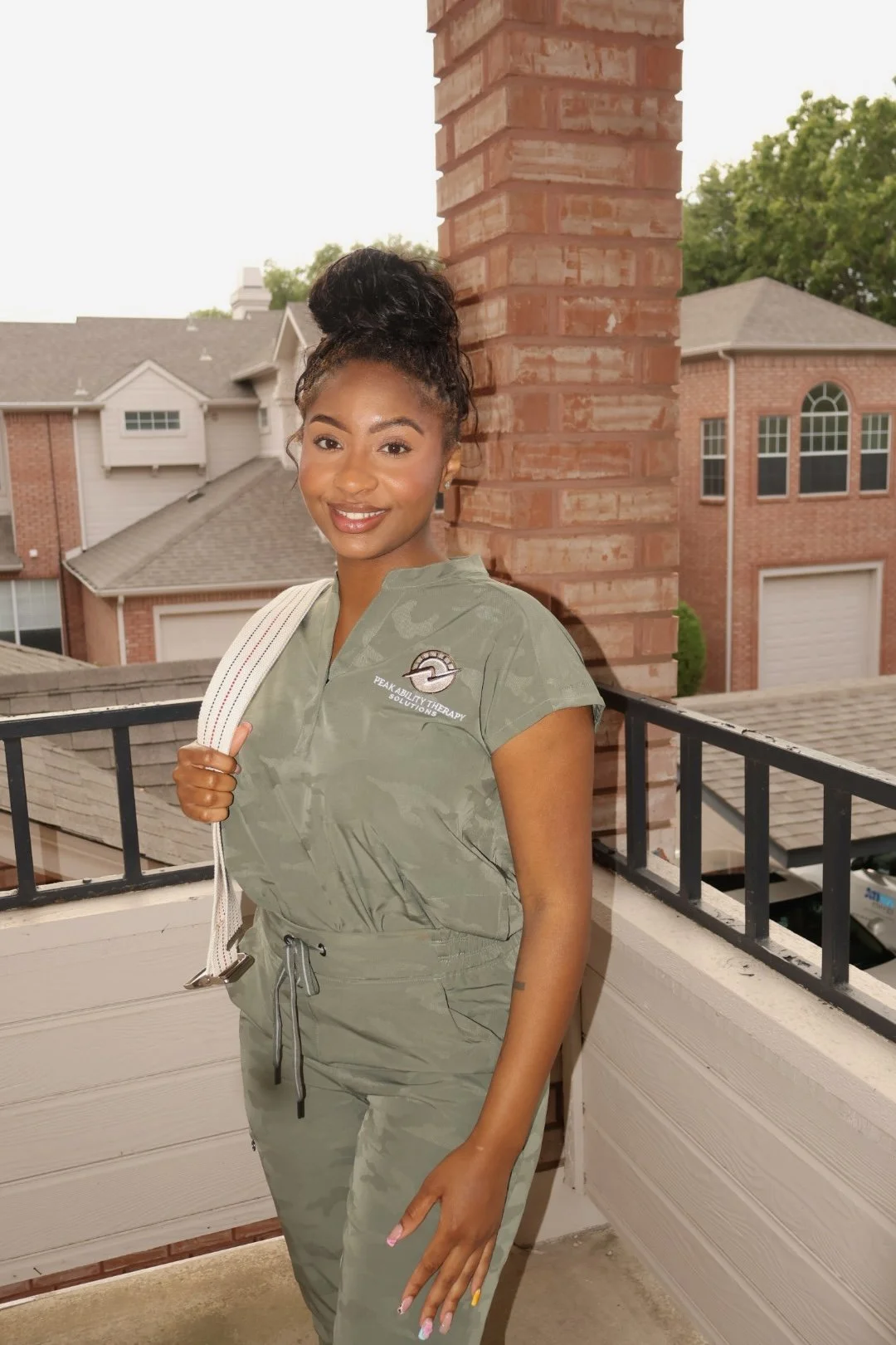 Smiling woman in green scrub uniform standing on a balcony with a view of houses and trees in the background.