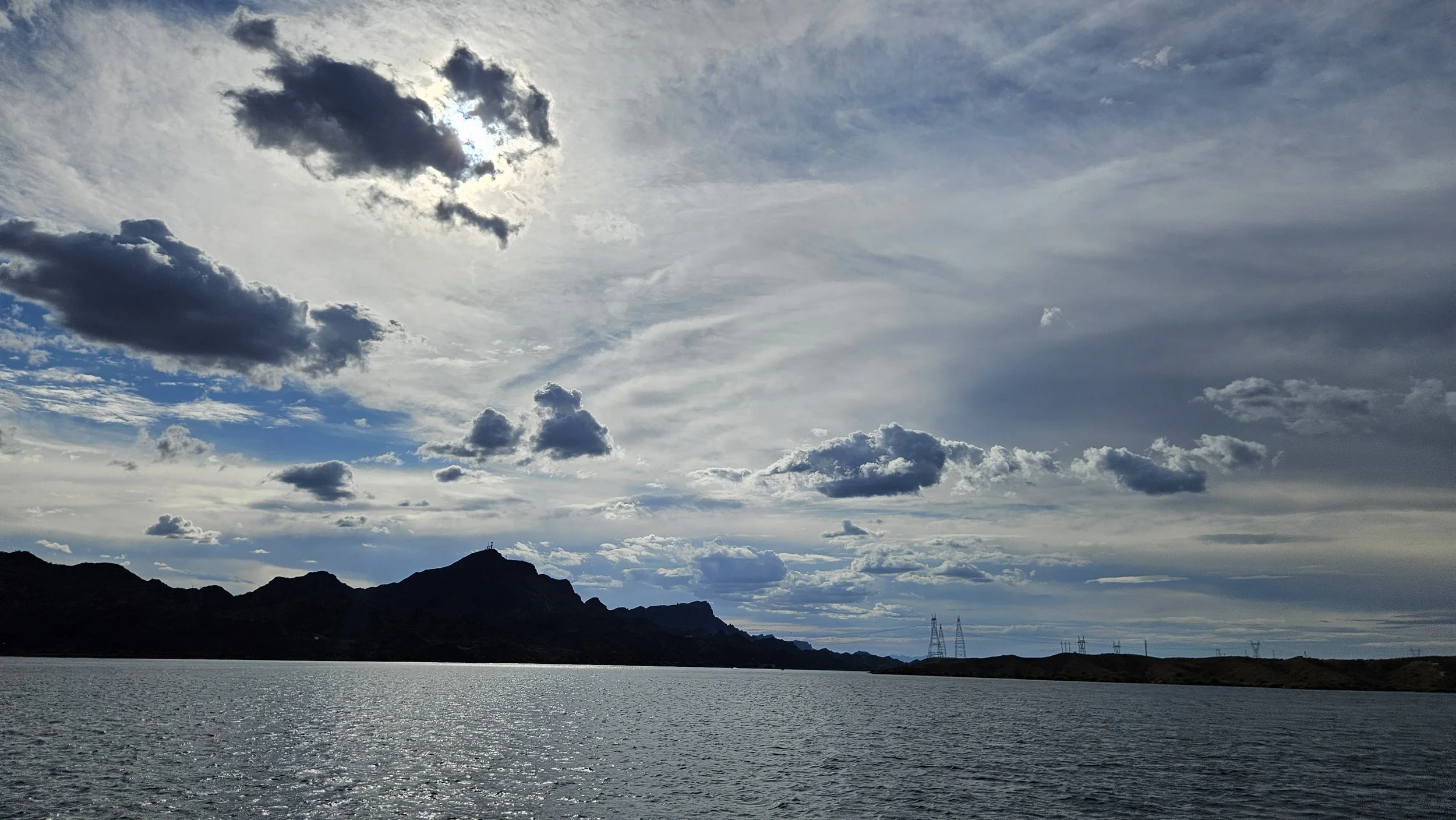 A scenic view of a body of water with mountains in the background under a partly cloudy sky with the sun peeking through the clouds.